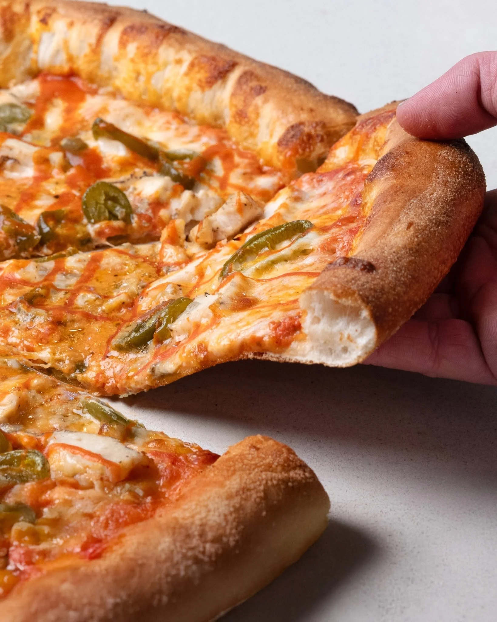 Close-up of a person holding a slice of pizza with melted cheese, green bell peppers, and tomato sauce on a white surface.