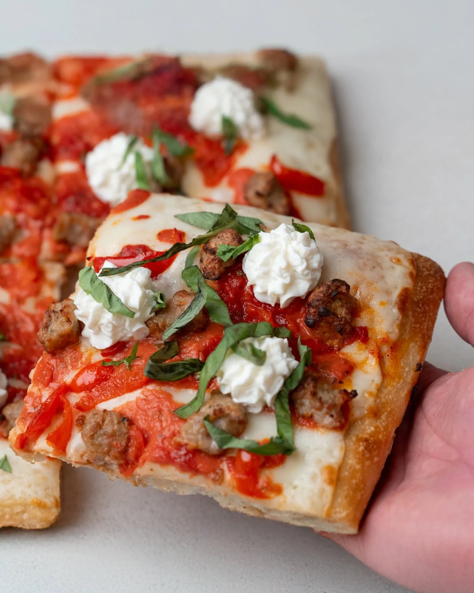 Close-up of a slice of pizza with pepperoni, sausage, tomato sauce, melted cheese, and dollops of ricotta cheese, garnished with basil, being held by a person's hand.
