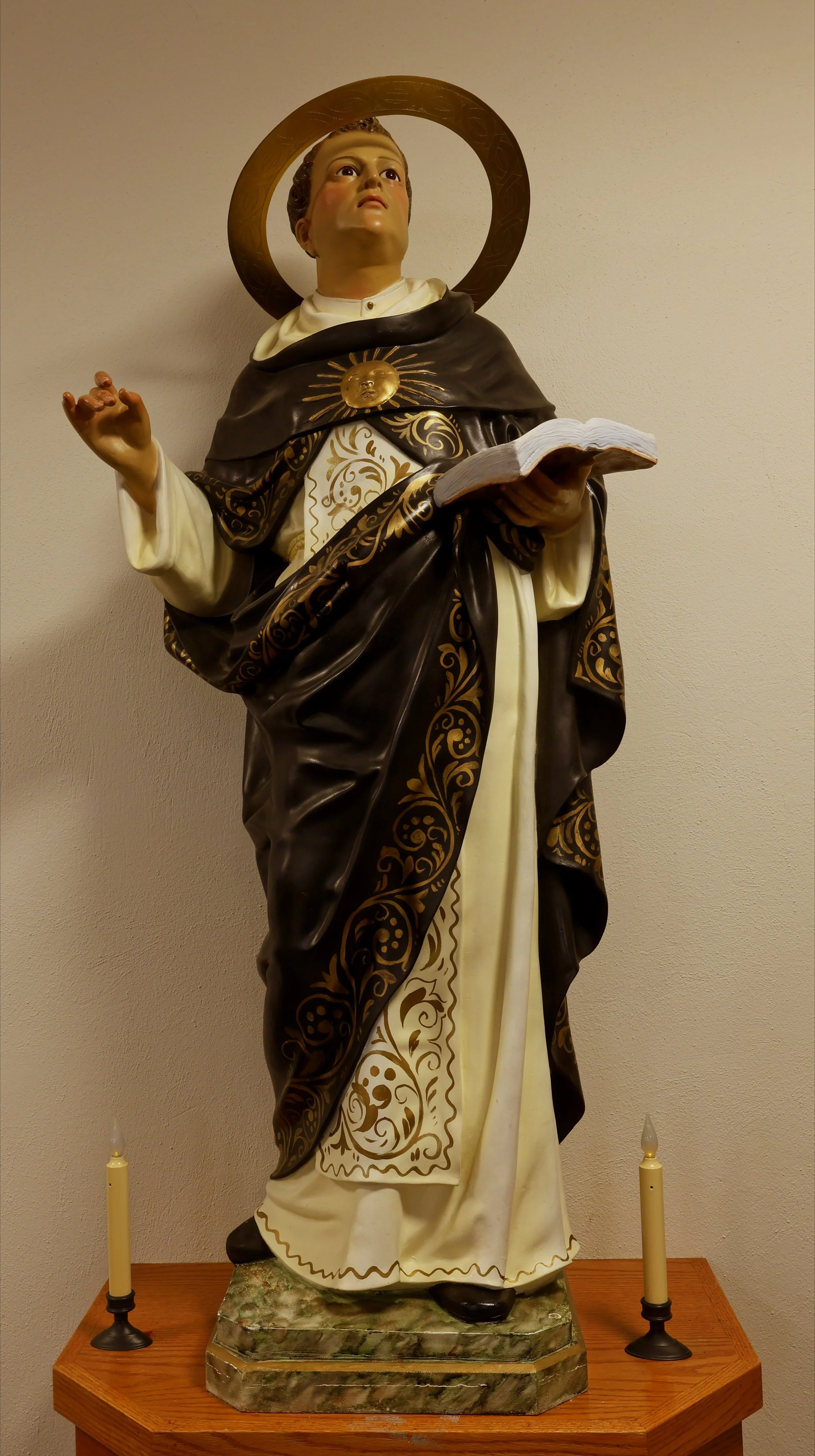 A religious statue of St Thomas Aquinas dressed in detailed robes with a halo around the head, holding a book, and standing on a marble base, flanked by two candles.