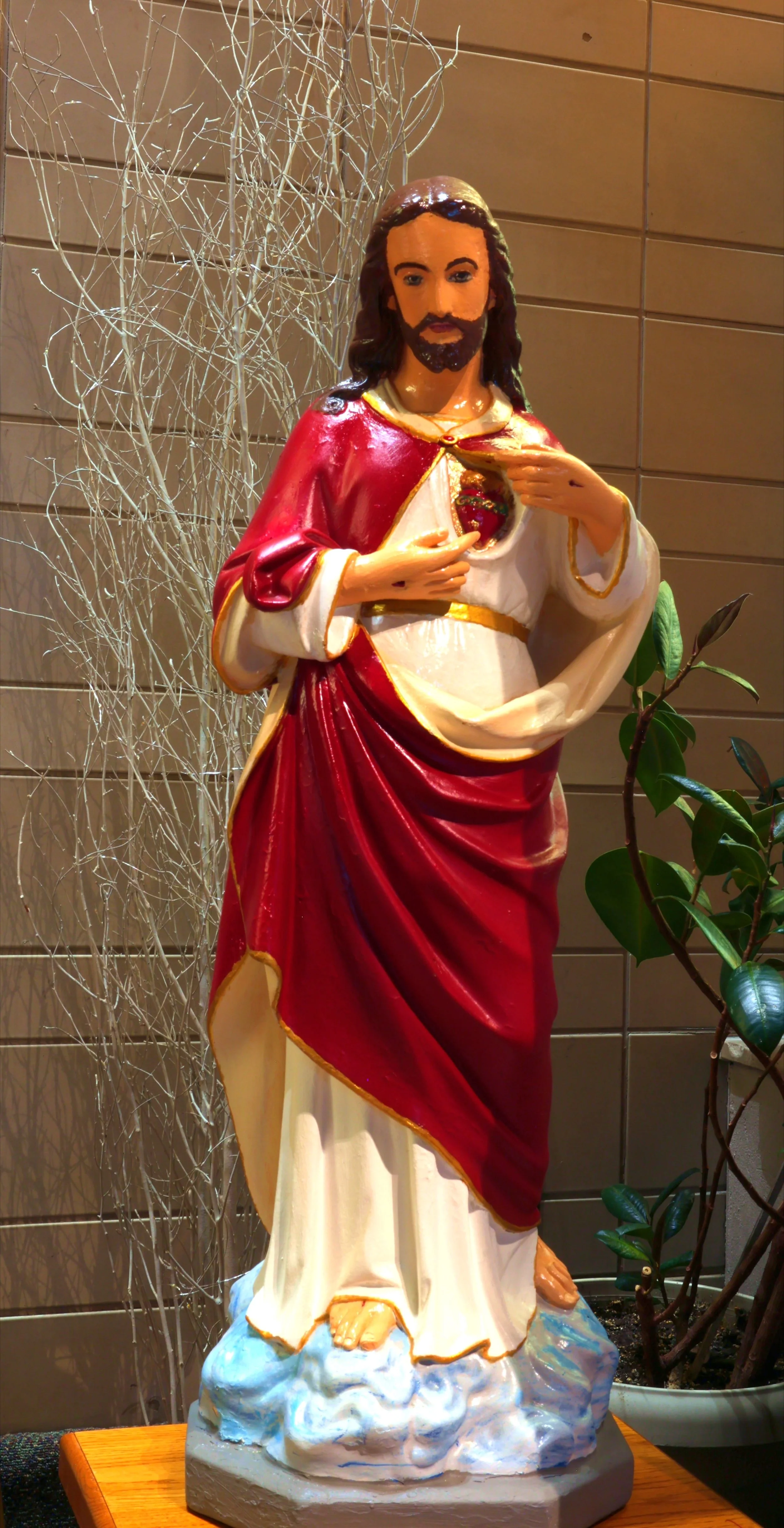 Colorful religious statue of Sacred Heart of Jesus, with a heart exposed on his chest, wearing a red and white robe, standing on a cloud base with a wooden surface and plants in the background.