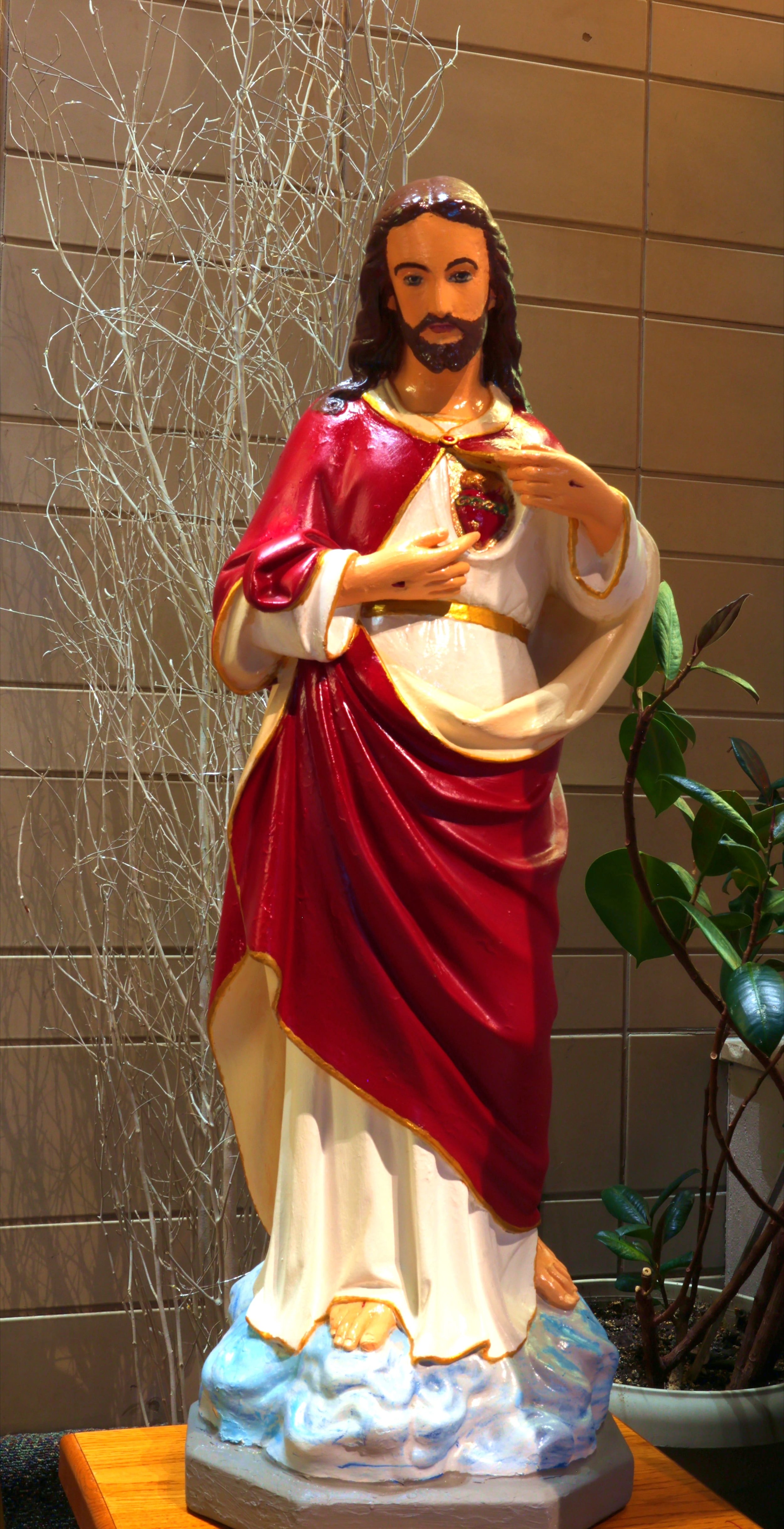 Colorful religious statue of Sacred Heart of Jesus, with a heart exposed on his chest, wearing a red and white robe, standing on a cloud base with a wooden surface and plants in the background.