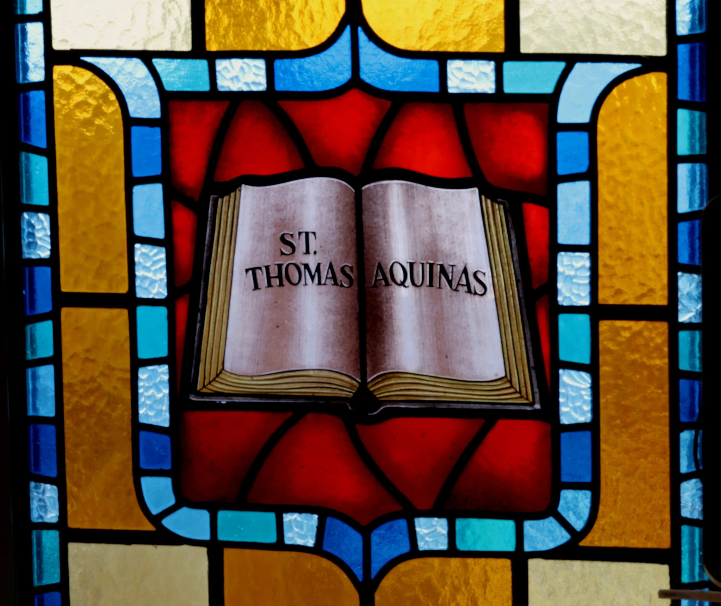 A stained glass window with the open book of Thomas Aquinas inscribed with his name. The window features geometric patterns in blue, yellow, and red.