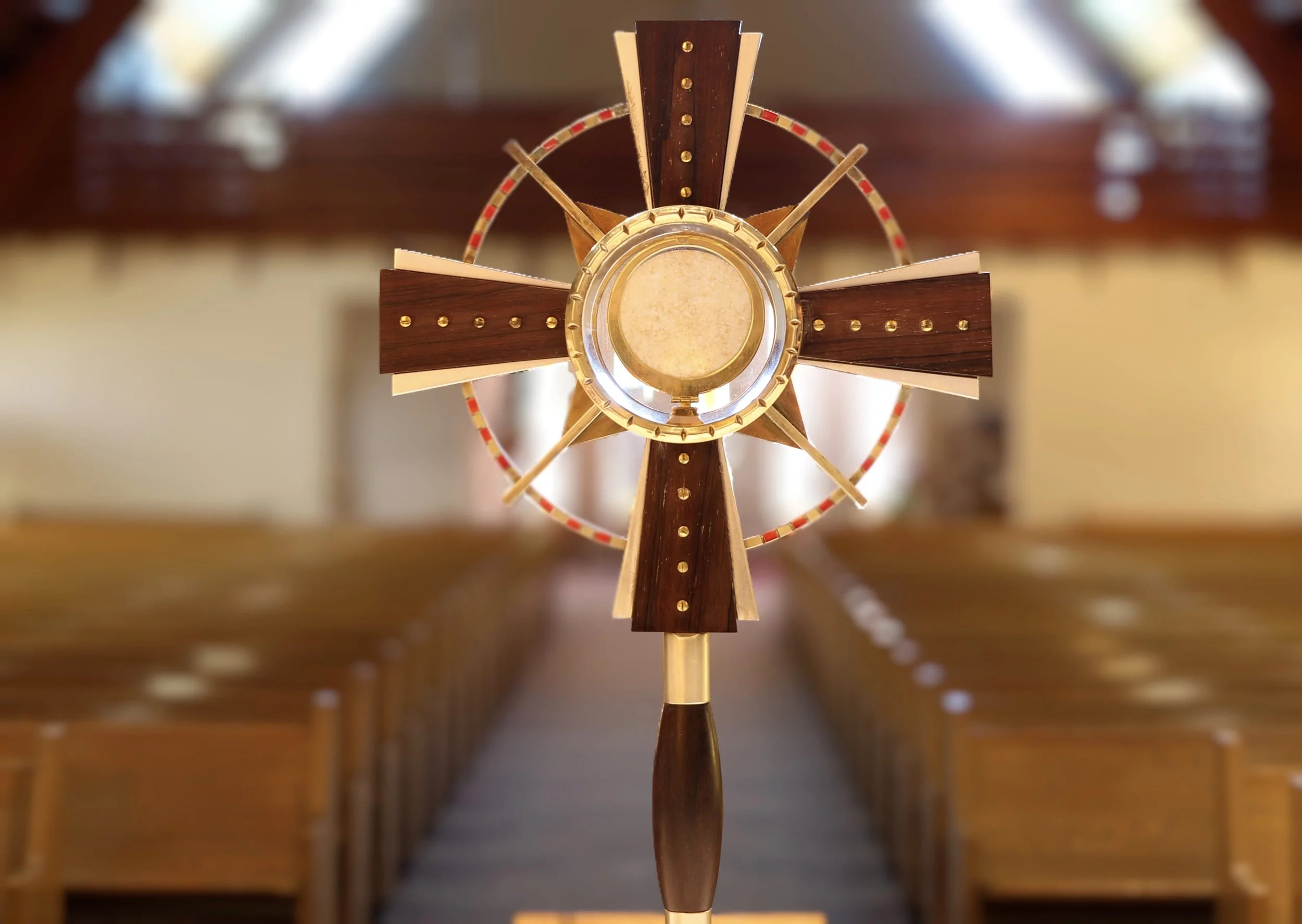A close-up view of a wooden monstrance with a host in the center, standing on a wooden base in a room with blurred background.