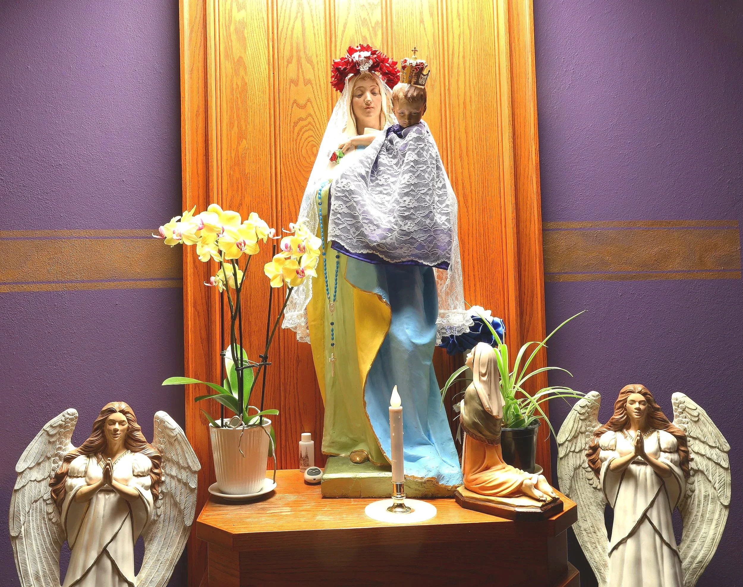 A religious display featuring a statue of the Virgin Mary holding the infant Jesus, both adorned with crowns and flowers, set against a purple wall with a wooden panel background. Surrounding the statues are angel figurines with wings, a potted orchid, and candles on a wooden table.