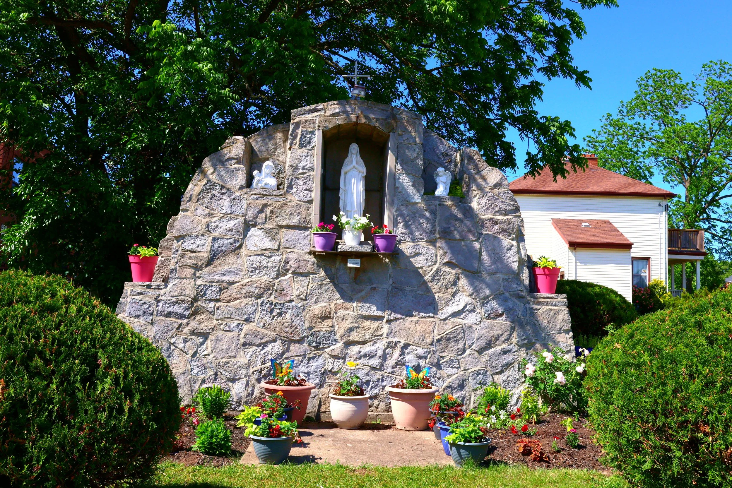 A stone Mary Grotto shrine with Marian statues in a garden, surrounded by colourful flower pots, bushes, and trees under a bright blue sky.
