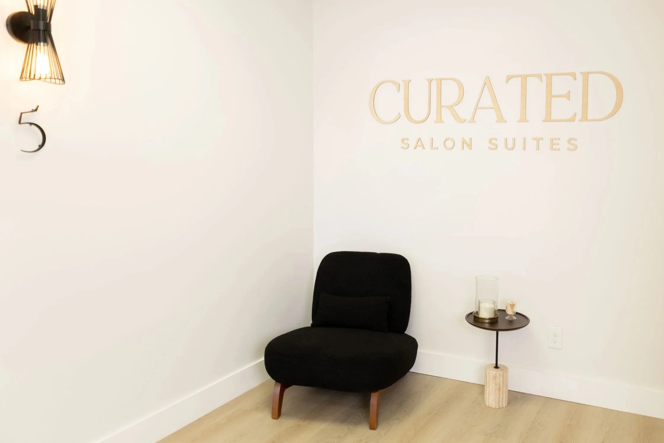 A minimalistic seating area with a black contemporary chair with wooden legs, beside a small round side table with decorative candles and glasses, against a plain white wall with the words 'CURED SALON SUITES' displayed in large light-colored letters