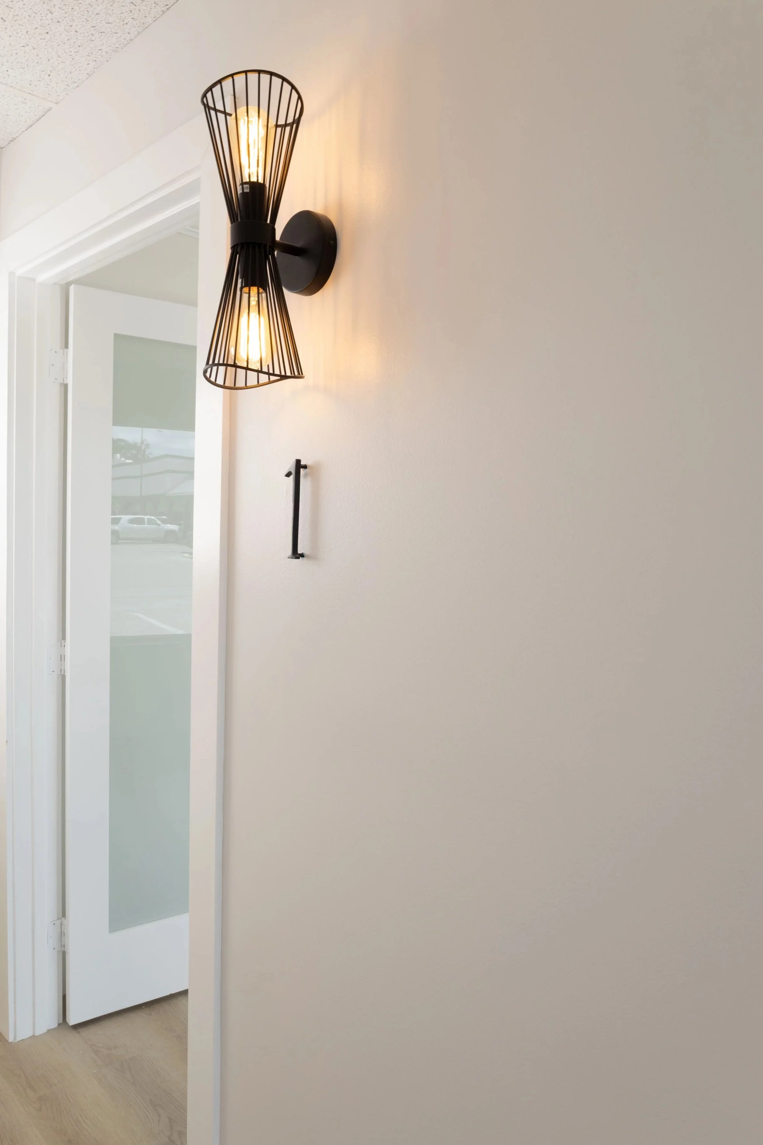 A modern wall sconce with two exposed filament light bulbs enclosed in black wireframe covers, mounted on a white wall, with a door and window showing a parking lot outside.
