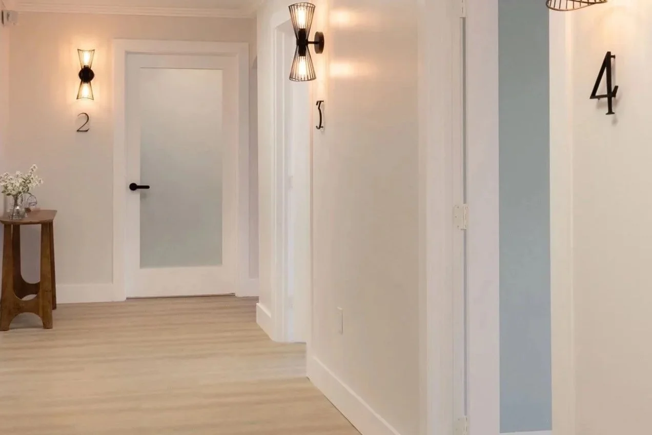A hallway with white walls and light wood flooring, featuring wall-mounted black and wire sconces, with the number 2 and letter J on the wall, and a door with frosted glass and a black handle.