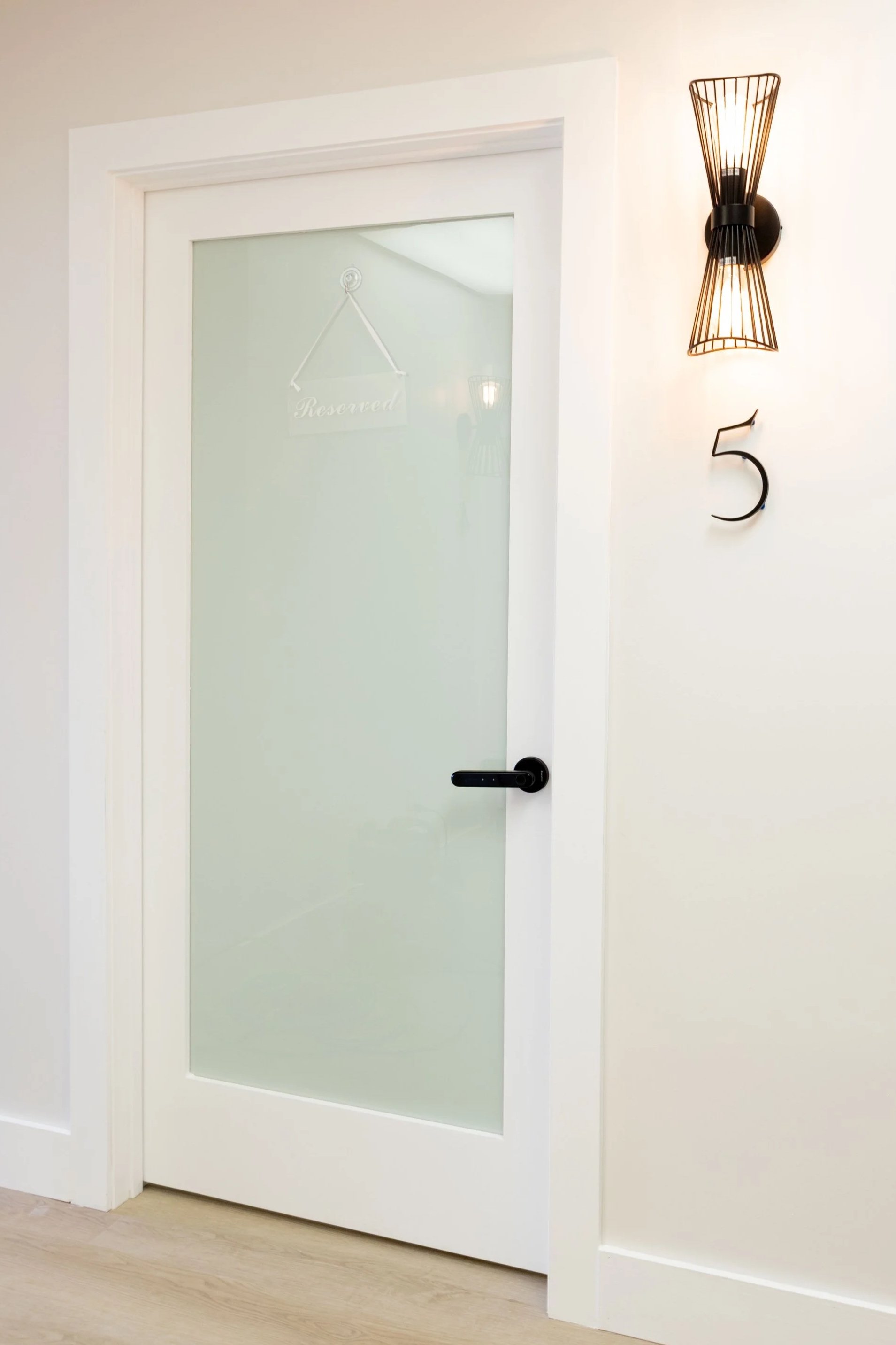 White door with frosted glass panel and black handle, mounted on a white wall, with a modern black wall sconce and a decorative black number five. private beauty salon suites in Zephyrhills