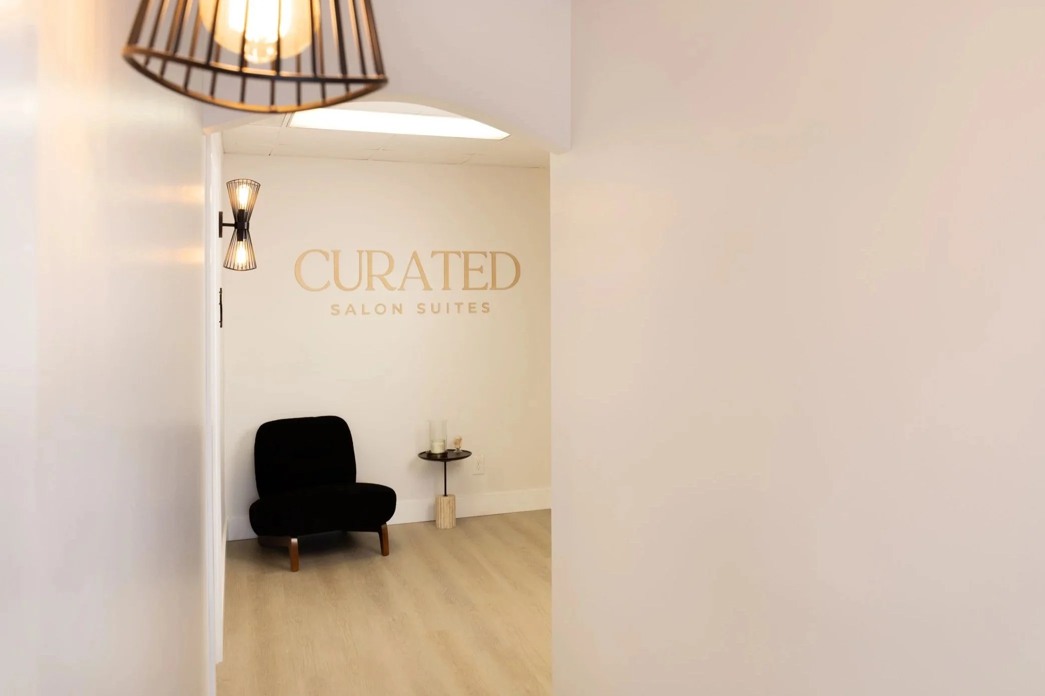 Interior of a salon suite with a wall displaying the sign 'Curated Salon Suites'. There is a black chair, a small side table with candles, and modern wall-mounted light fixtures. The space has a minimalist and elegant design with neutral colors.