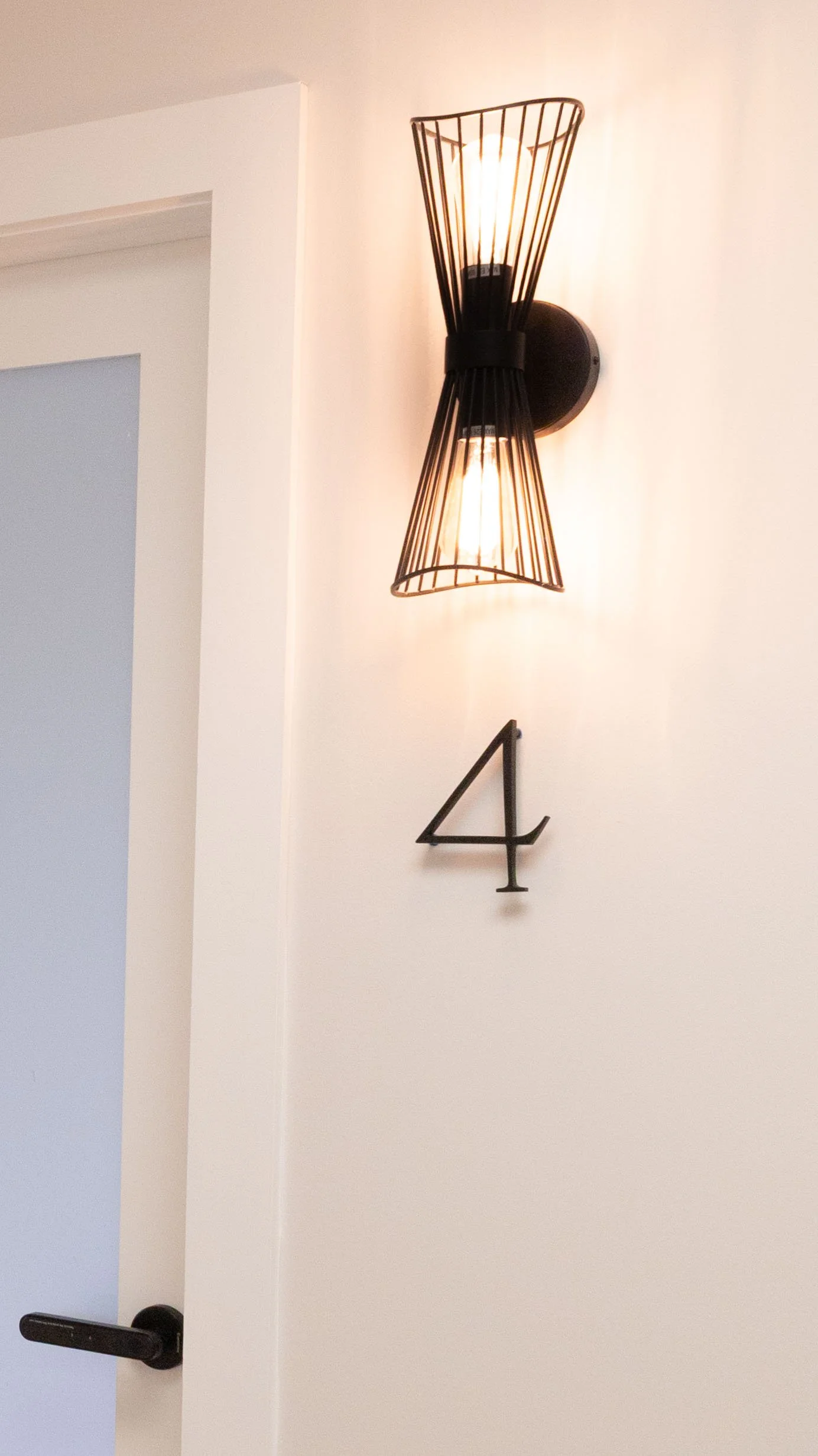 Wall-mounted light fixture with exposed bulbs and black wireframe cover, beside a door with a black handle, and black wall art displaying the number 4. Zephyrhills salon suites available for lease