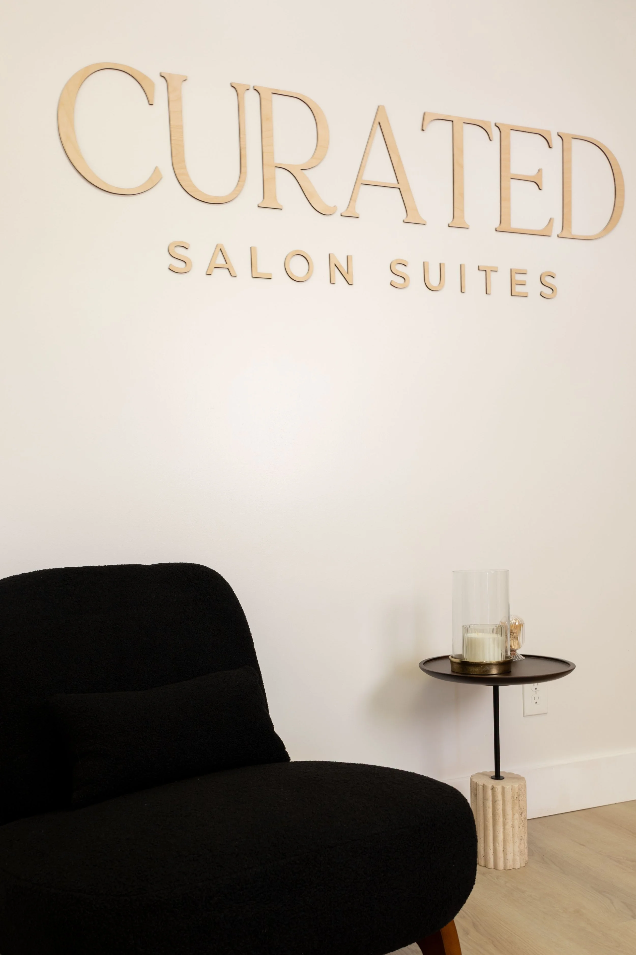Interior of a modern living space with a black couch, a side table with candles and a tall glass, and a wall sign reading 'CURED SALON SUITES'.
