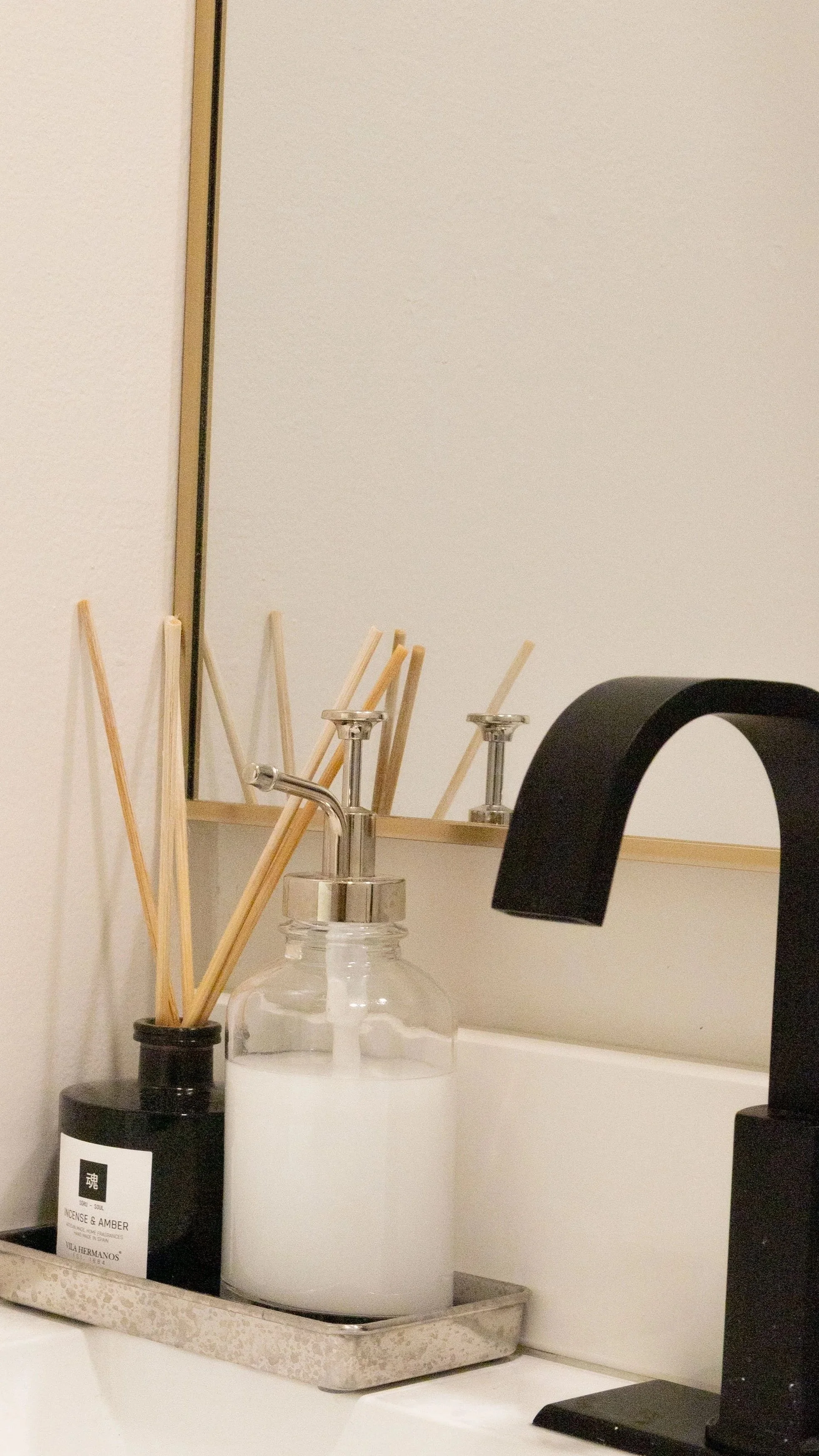 Countertop with reed diffusers, a soap dispenser, and a black faucet, reflected in a mirror.