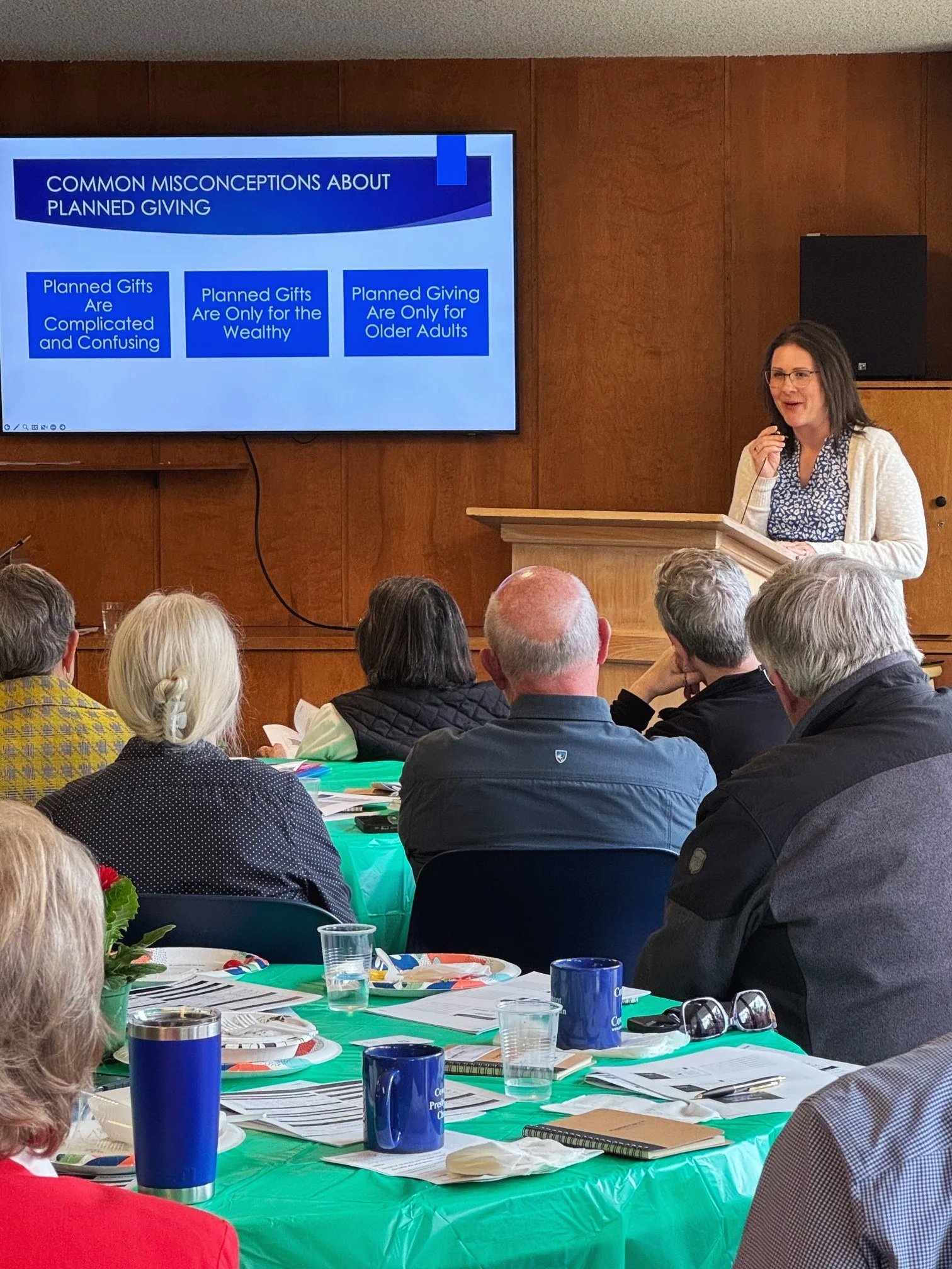 A woman standing at a podium speaking to an audience during a presentation titled 'Common Misconceptions About Planned Giving.' The audience, seated around a large table with papers, notebooks, and blue mugs, listens attentively in a wood-paneled room.
