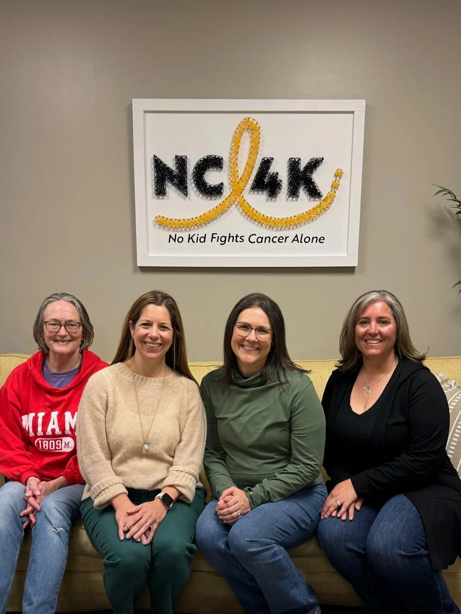 Four women sitting on a couch in front of a wall with a sign that reads 'NC4K' and 'No Kid Fights Cancer Alone.'