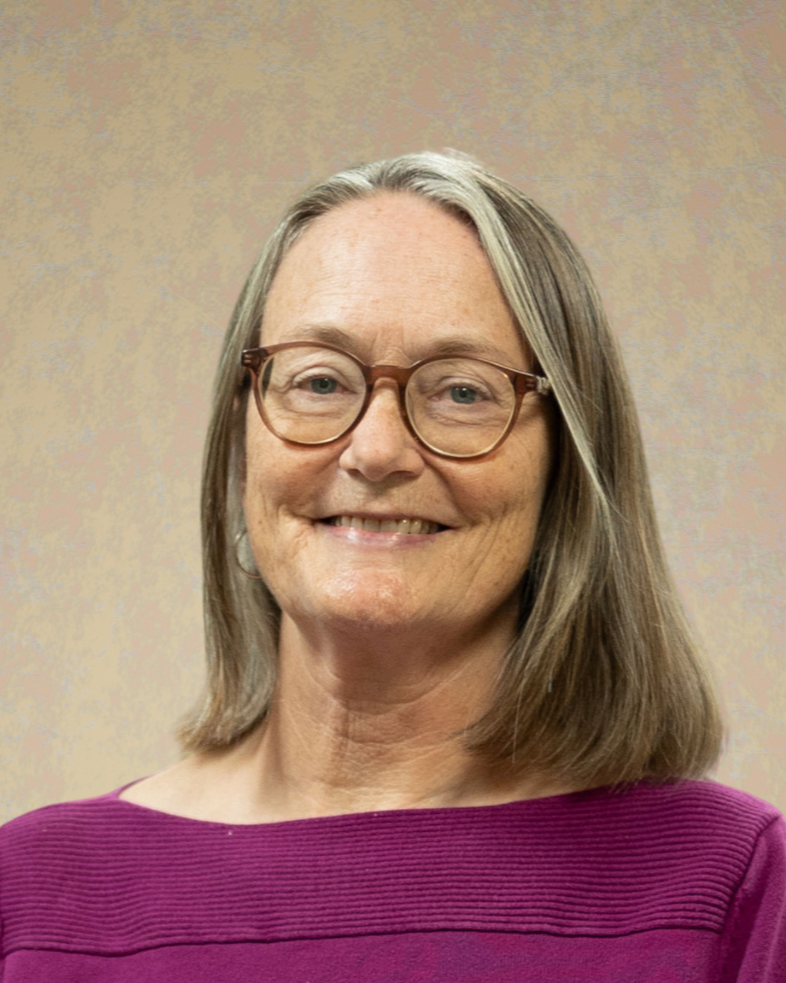 A woman with shoulder-length gray hair and glasses, smiling, dressed in a purple top, against a beige background.