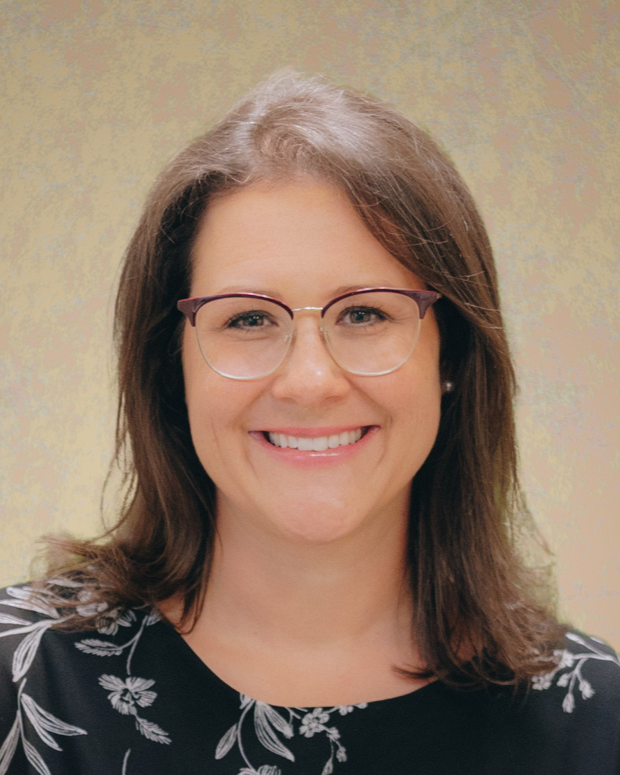 A woman with glasses and shoulder-length brown hair smiling in front of a plain beige background.