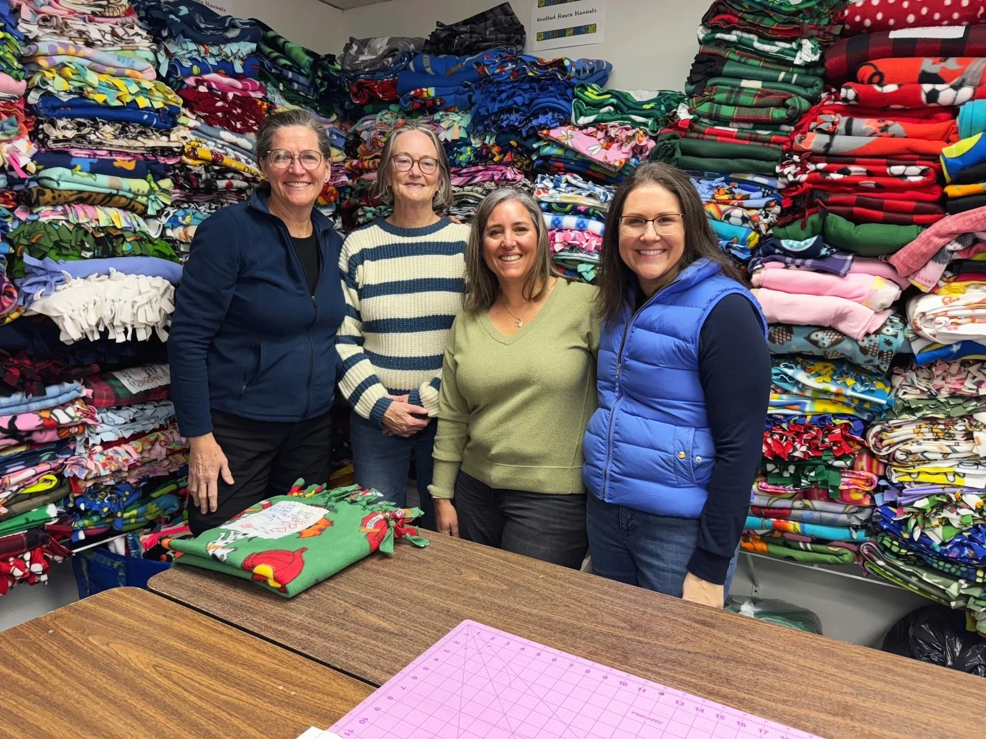 Four women standing behind a table filled with folded Christmas-themed fleece blankets at a donation or charity event, with shelves of colorful fleece blankets in the background.