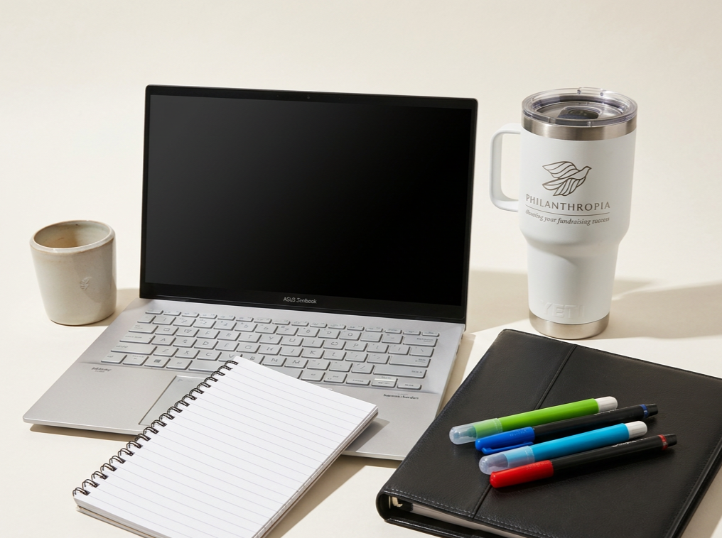 A workspace with a laptop, a spiral notebook, a black portfolio, four colorful pens, a mug, and a travel tumbler on a white surface.