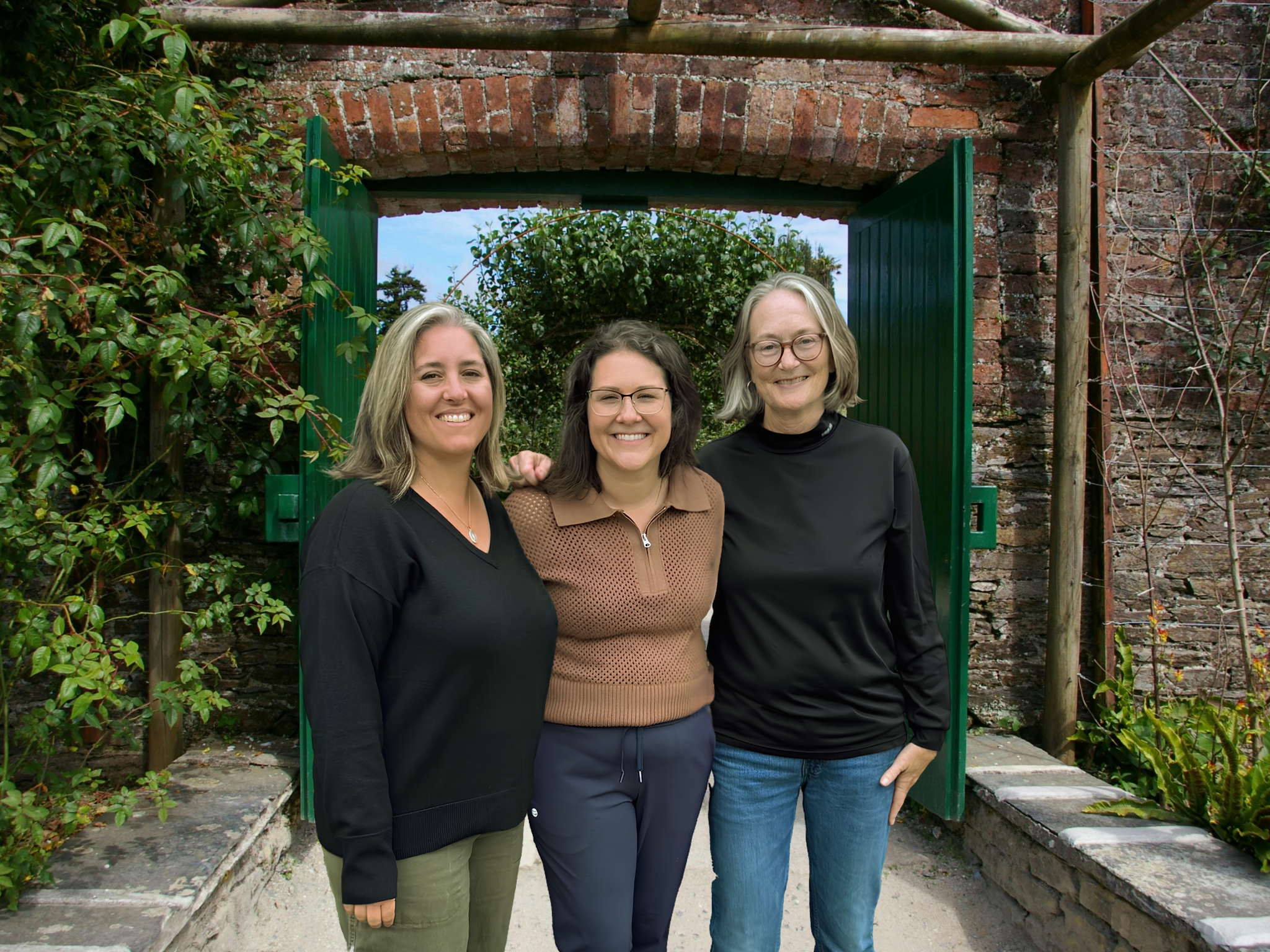 Three women standing together outdoors, smiling, in front of an archway with green shutters and brick wall, with greenery surrounding.