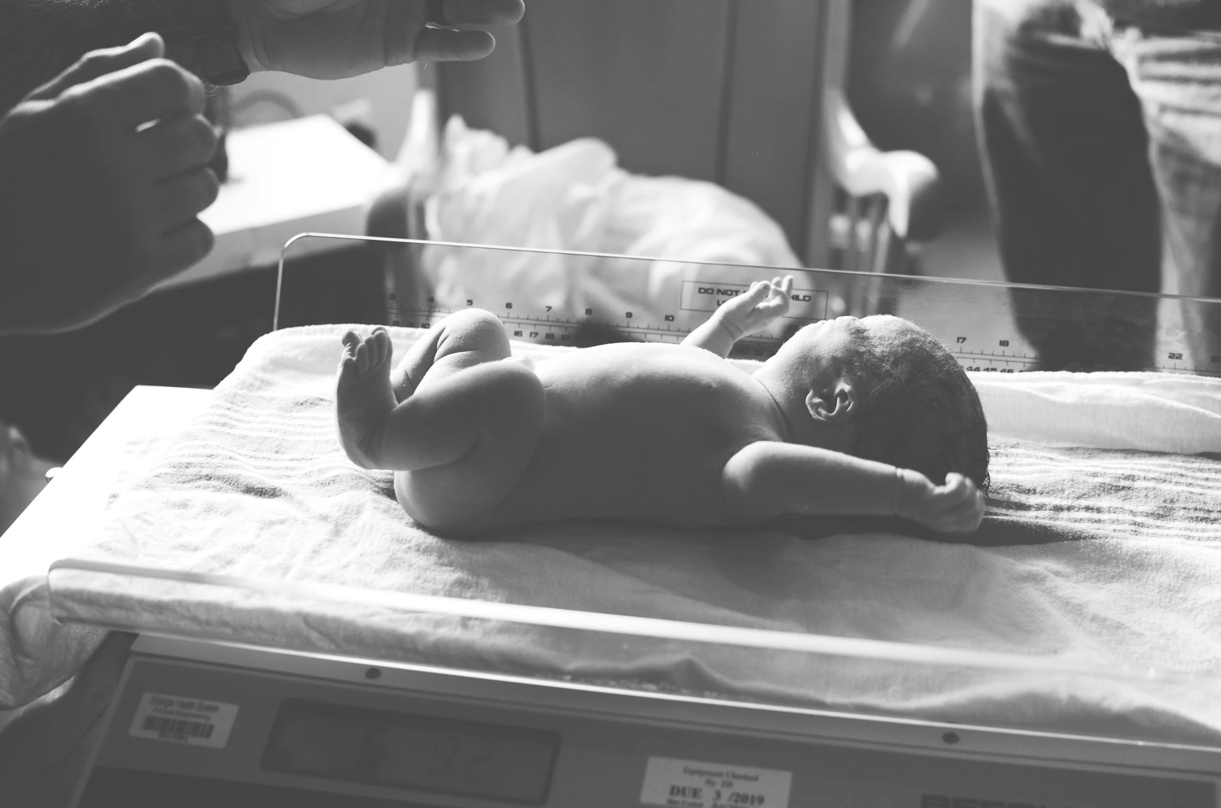 Black and white photo of a newborn baby lying on a hospital bed with a measuring scale beneath. A caregiver's hand reaches over the baby, and a clear plastic barrier with a warning sign is in the foreground.