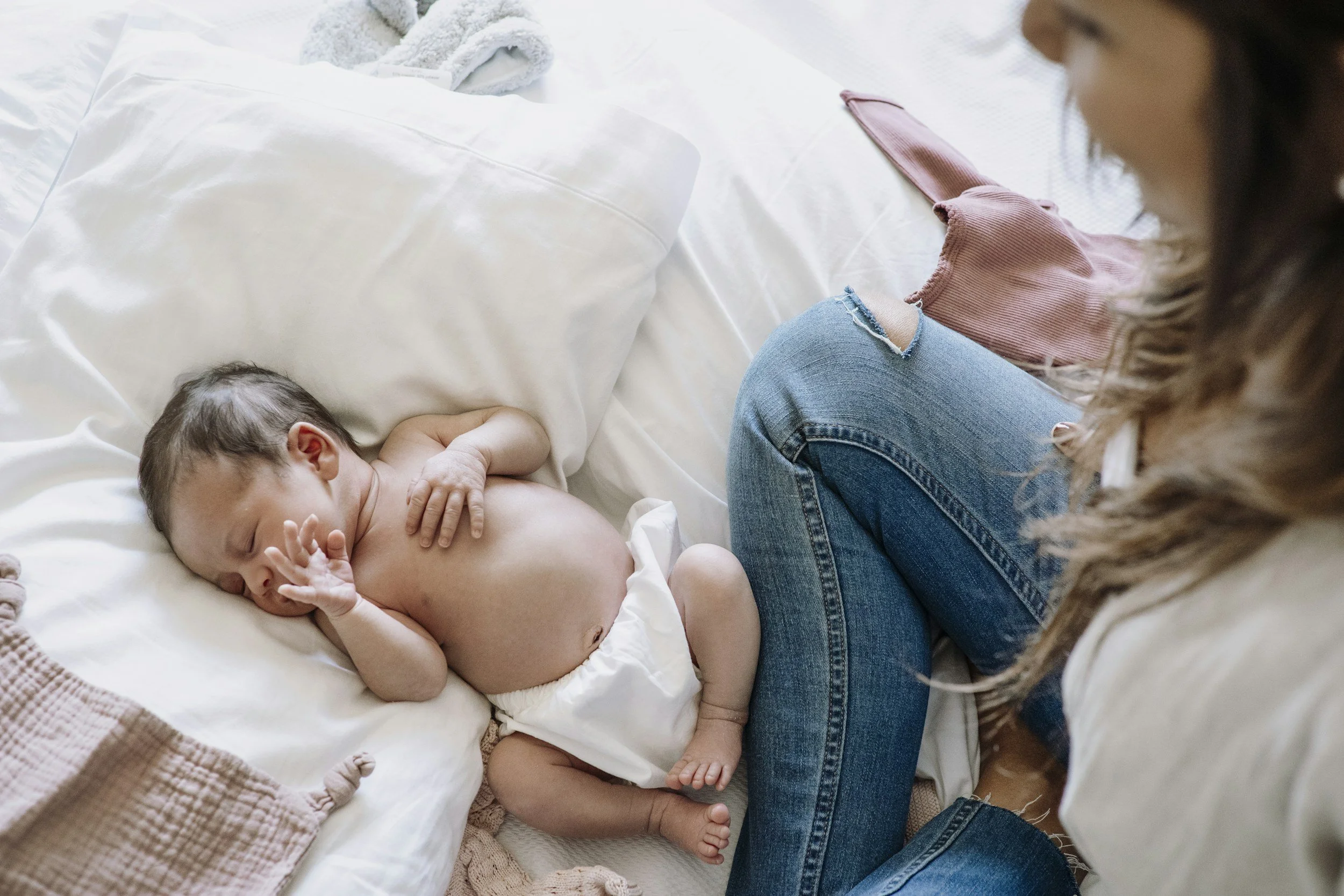 A woman with curly hair and ripped jeans sitting on a bed, looking at a sleeping baby lying on a pillow and blanket. The baby is topless, sleeping peacefully, with one hand near the face.