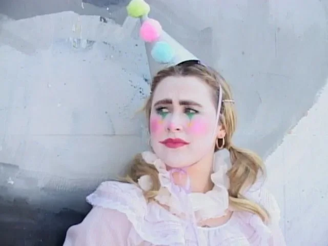 A woman dressed as a clown with colorful blush and eye makeup, wearing a pastel party hat with pom-poms, leaning against a wall with a distressed expression.