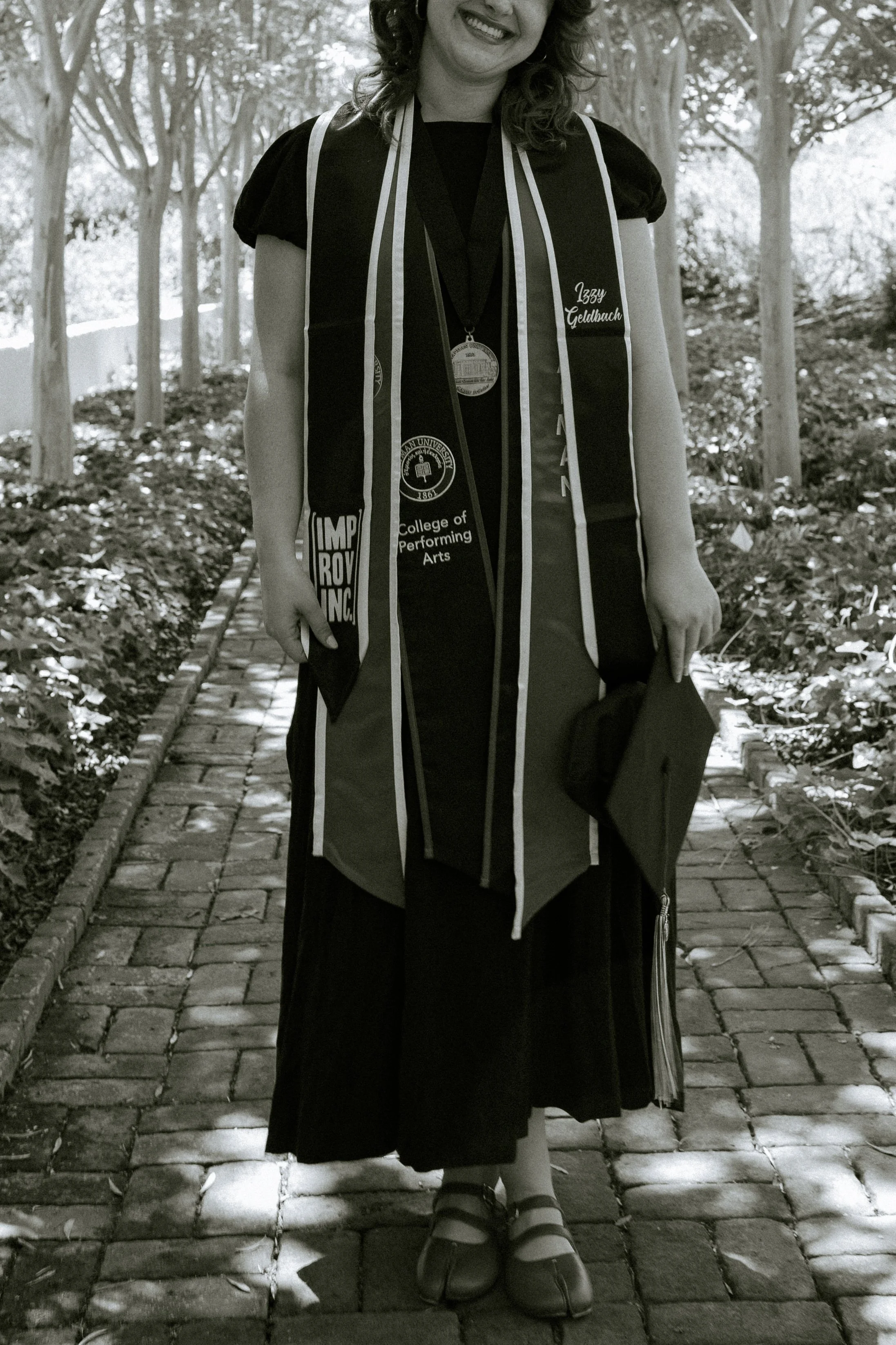 Woman wearing a graduation gown and cap, standing on a brick pathway surrounded by trees.