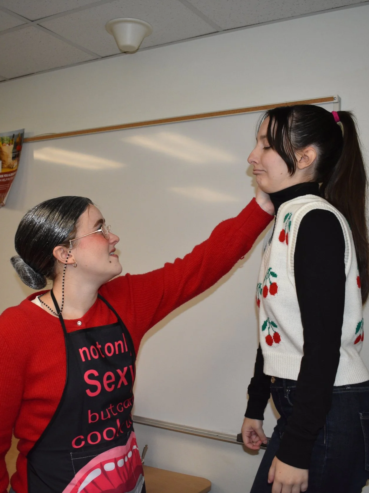 A woman with dark hair, glasses, and earrings, wearing a black apron with red and white text, touching the chin of a young woman with dark hair in a ponytail, wearing a black turtleneck and a white sleeveless sweater decorated with cherries, in a room with white walls and ceiling.