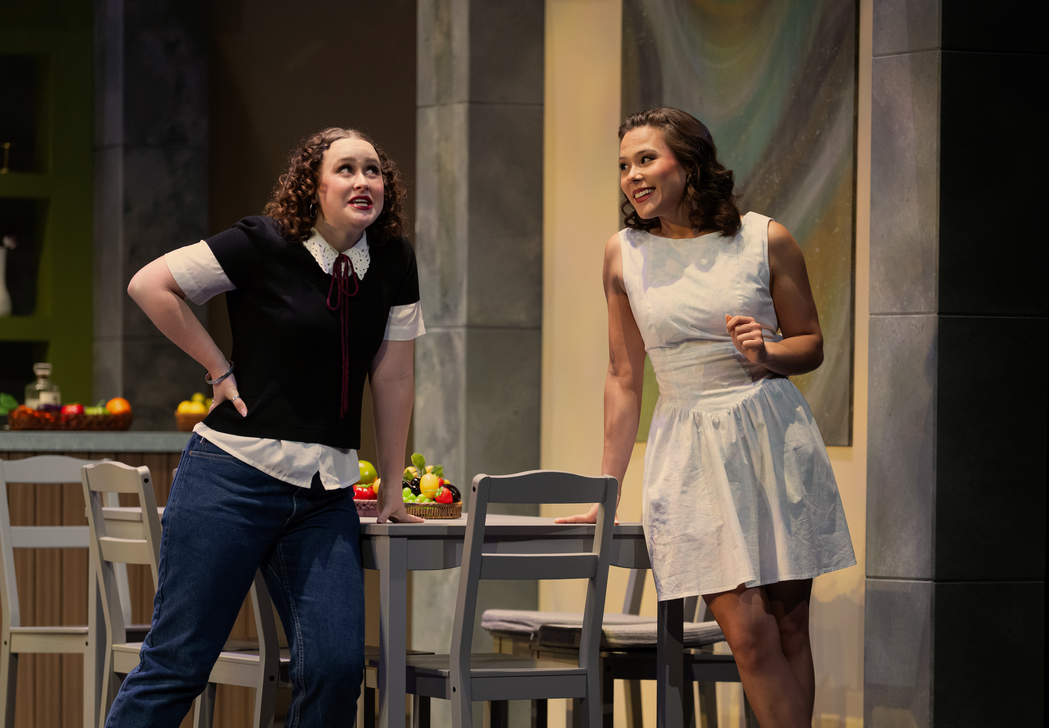 Two women having a conversation in a modern kitchen set. One woman is dressed casually with a black top and jeans, the other in a white dress. There are fruits and vegetables on the table.
