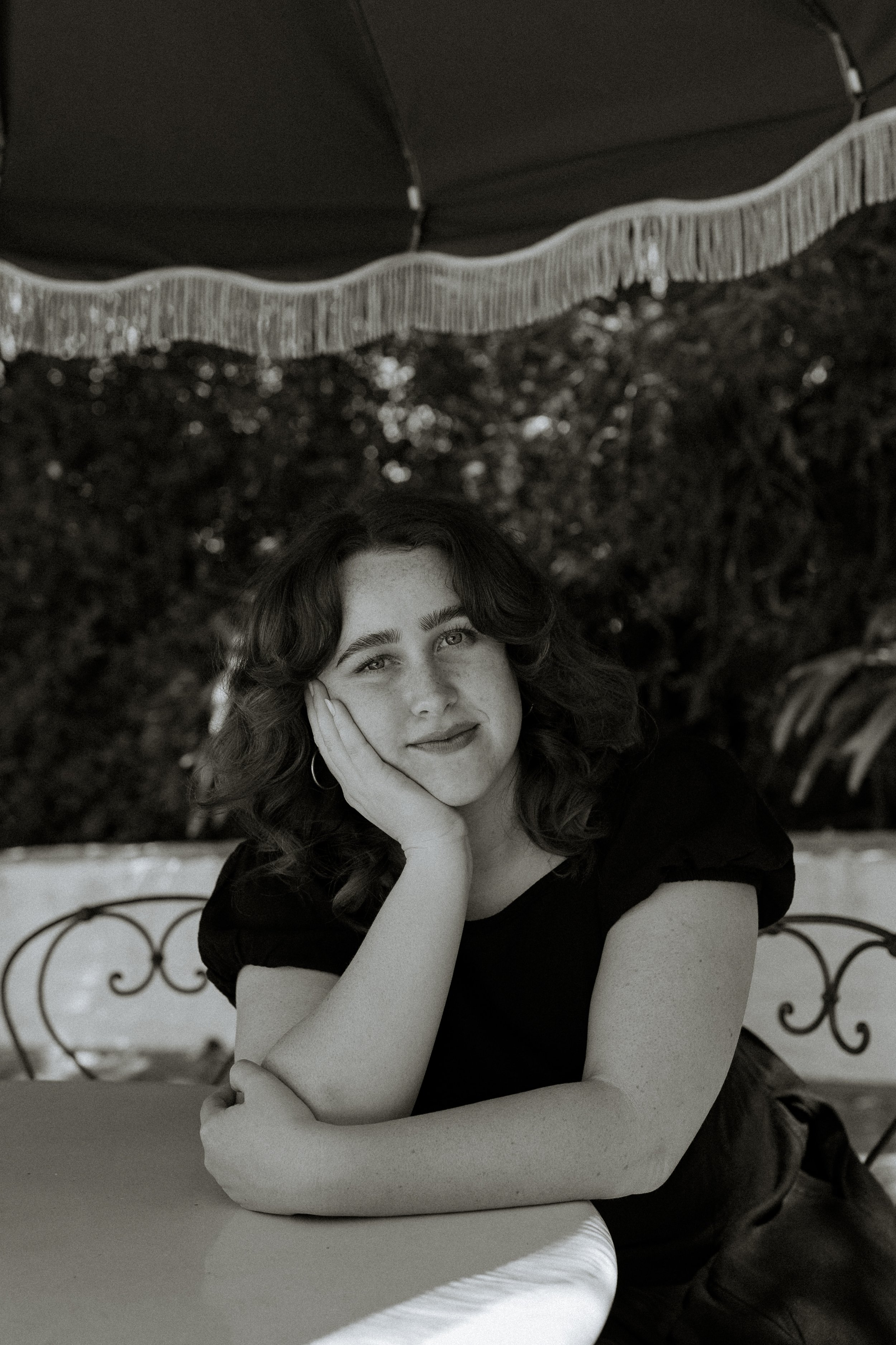 Black and white portrait of a woman with curly hair, sitting at a table with a metal decorative headboard behind her, resting her face on her hand and looking at the camera.