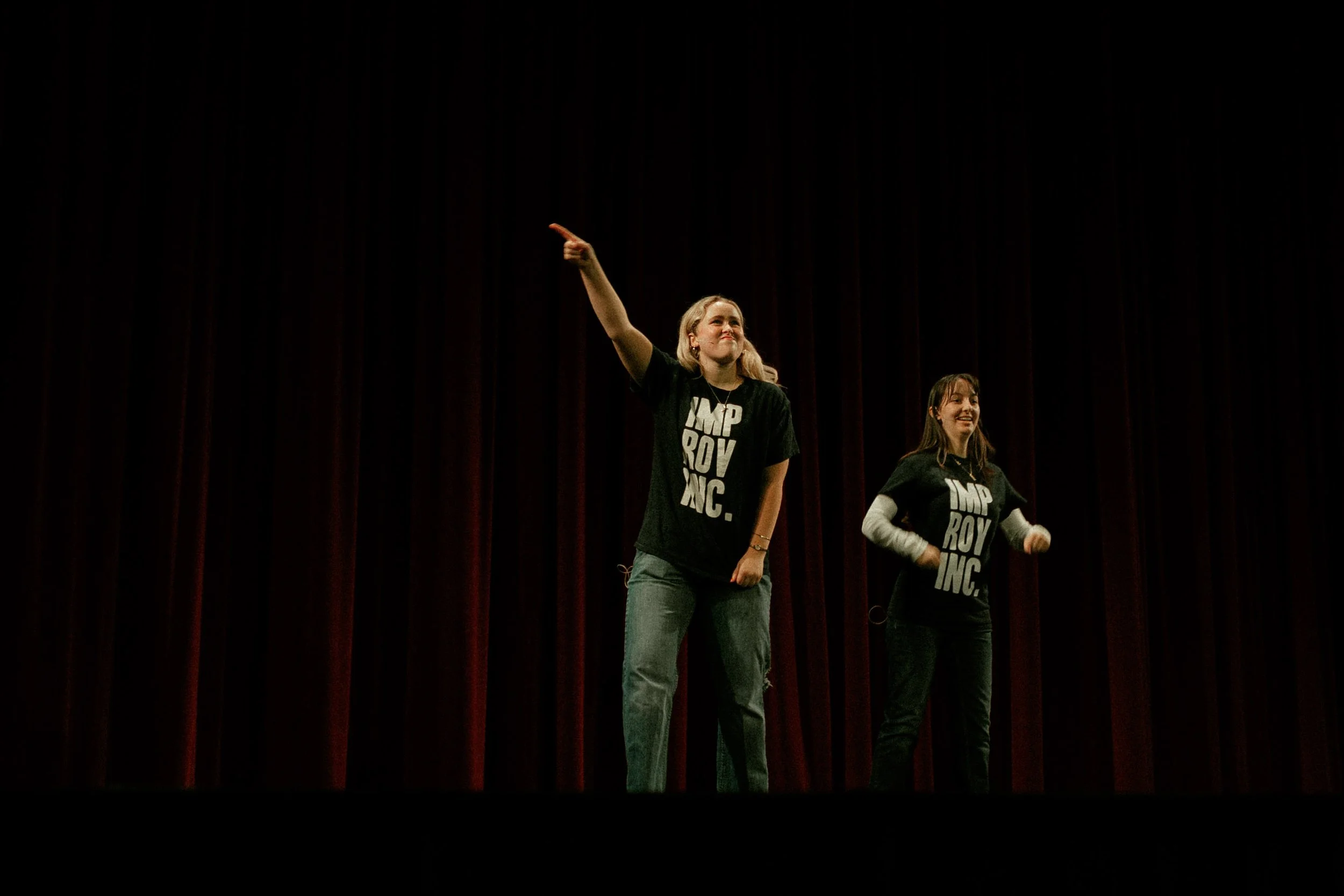 Two women on stage, wearing black t-shirts with bold white text, standing in front of closed red stage curtains. One woman is pointing upward and smiling, while the other is smiling with her hands on her hips.