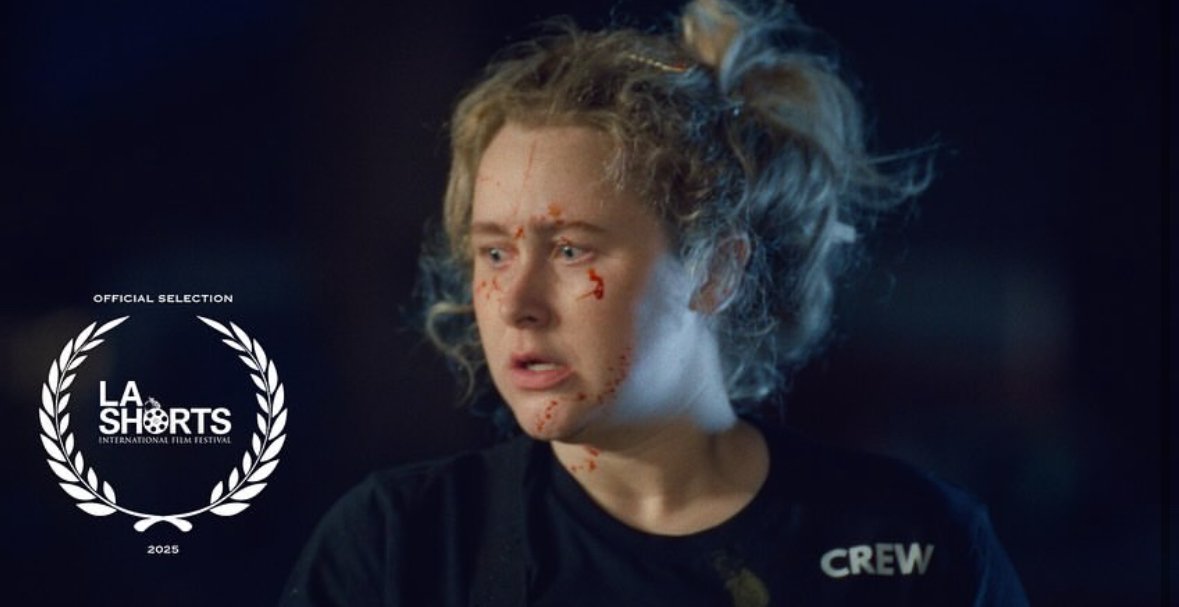 A young woman with blonde curly hair tied in a loose bun, wearing a black shirt with 'CREW' written on it, has cuts and bruises on her face and appears distressed. The background is dark, and there is a La Shorts 2025 international film festival logo on the left side of the image.