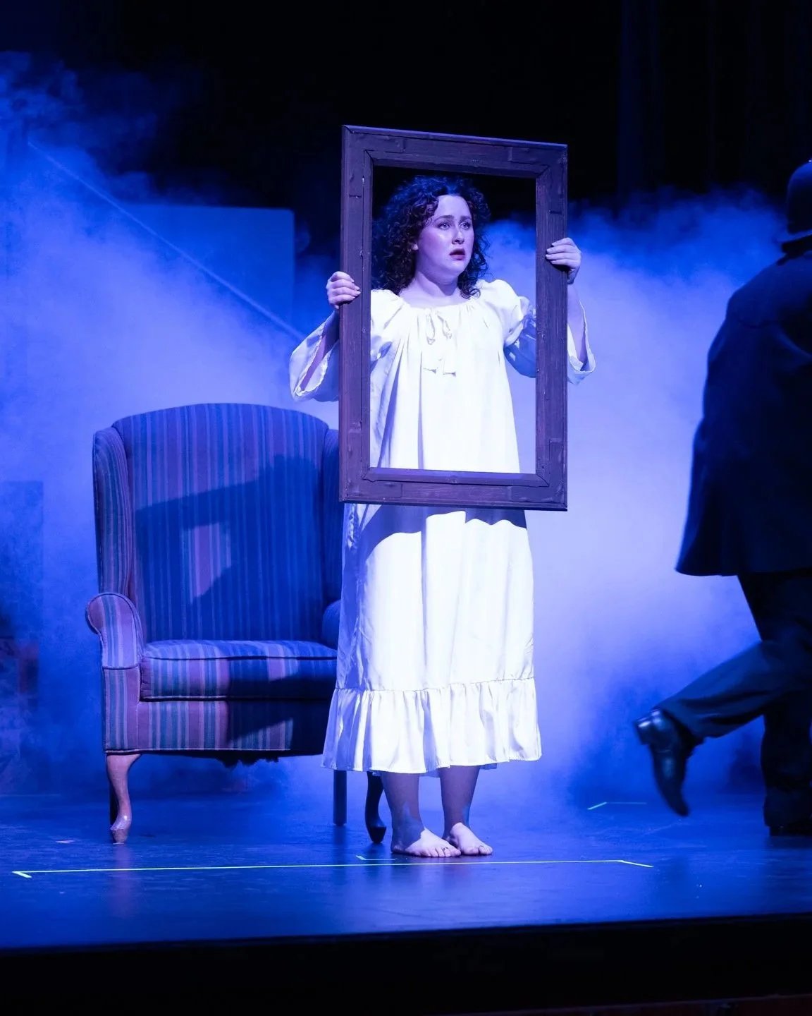 A woman dressed in a nightgown holding a picture frame in front of her, standing barefoot on stage during a theatrical performance with smoky blue lighting, an armchair, and a person partially visible to the right.