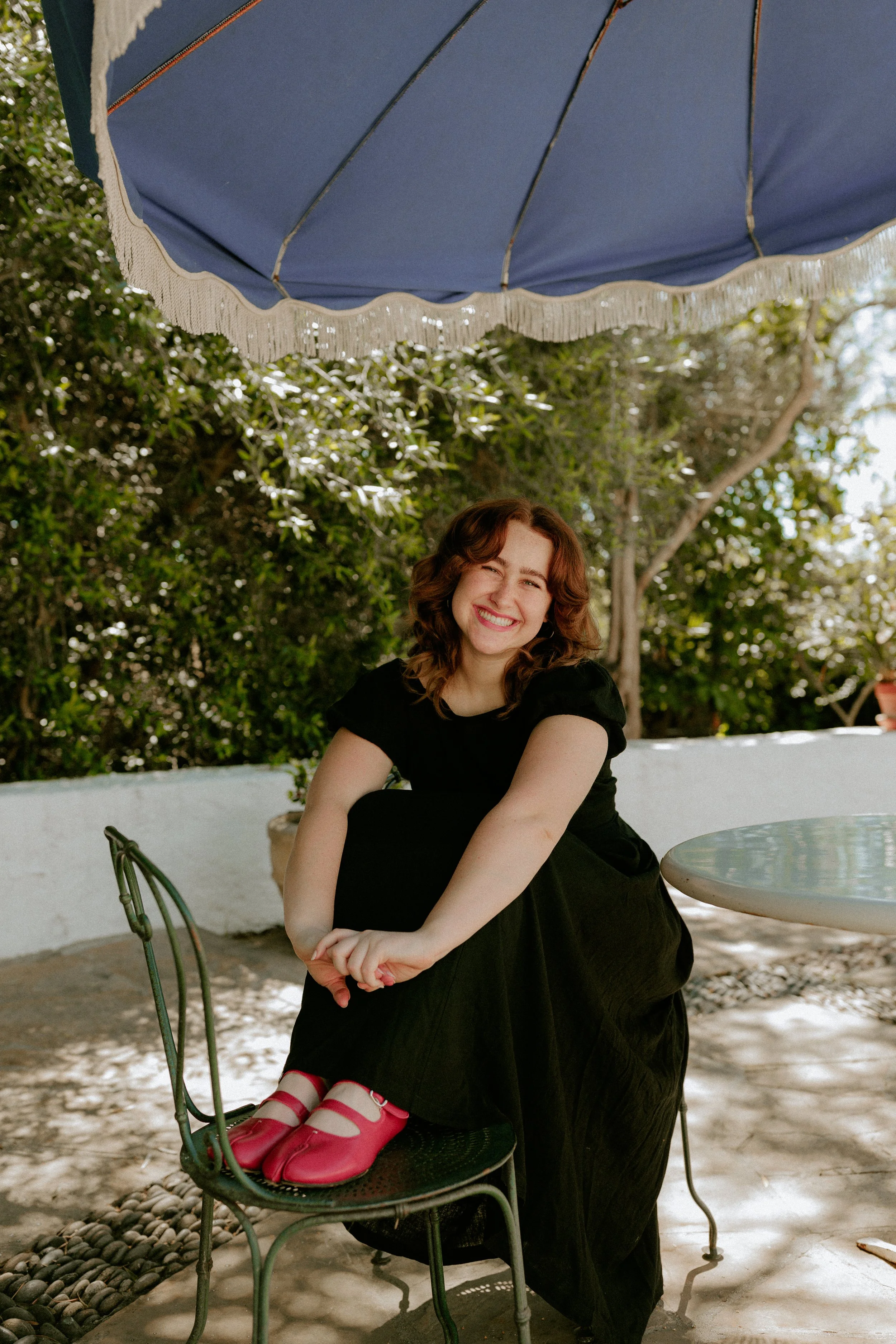 A young woman with curly brown hair and light skin is sitting under a blue patio umbrella outdoors. She is smiling widely with one eye closed, wearing a black dress and red shoes. She is seated on a green metal chair at a round marble table with a white wall behind her and shrubs in the background.