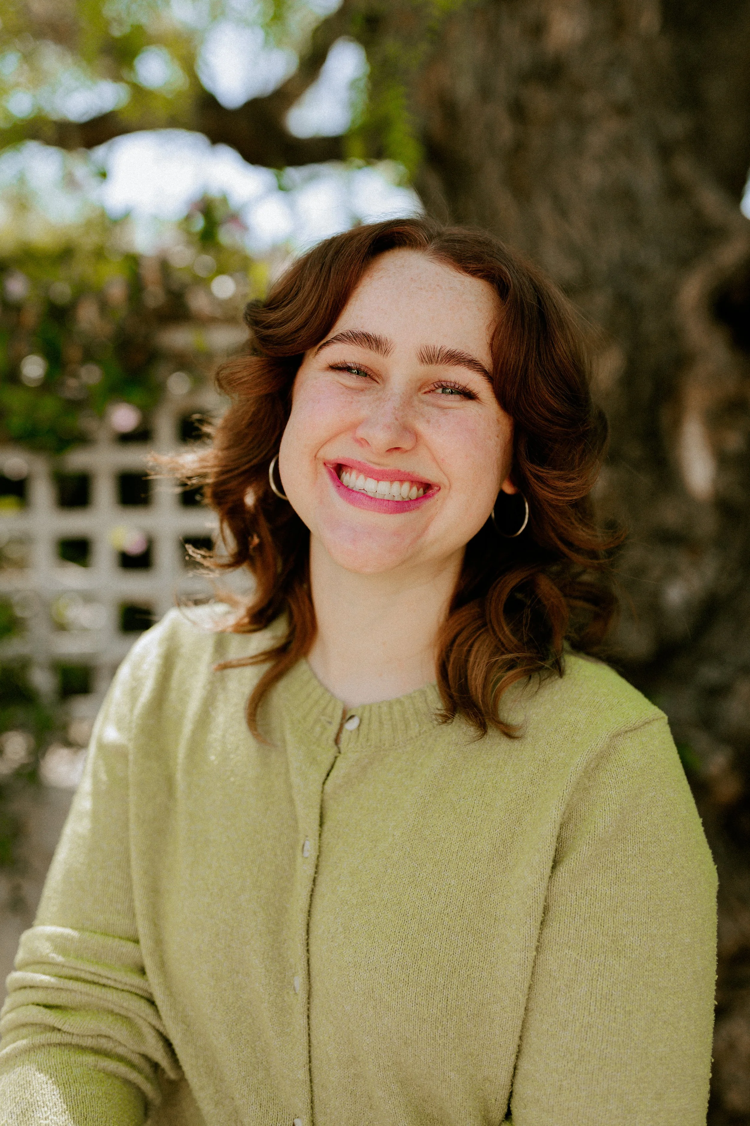 A woman with brown hair and fair skin smiling outdoors in front of a tree and a white picket fence, wearing a light green cardigan.