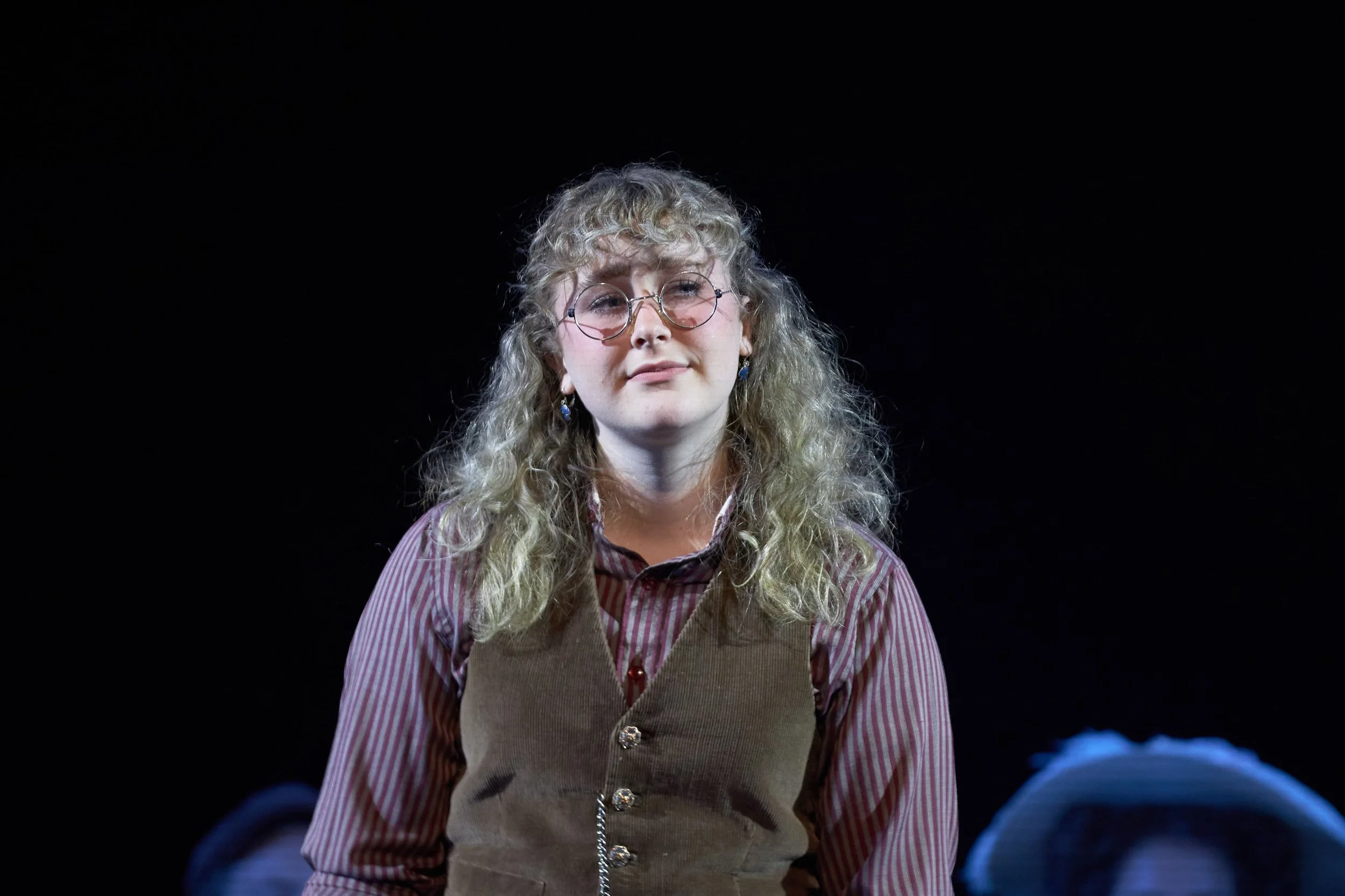 A young woman with curly blonde hair, glasses, earrings, and a striped shirt, sitting or standing against a black background.