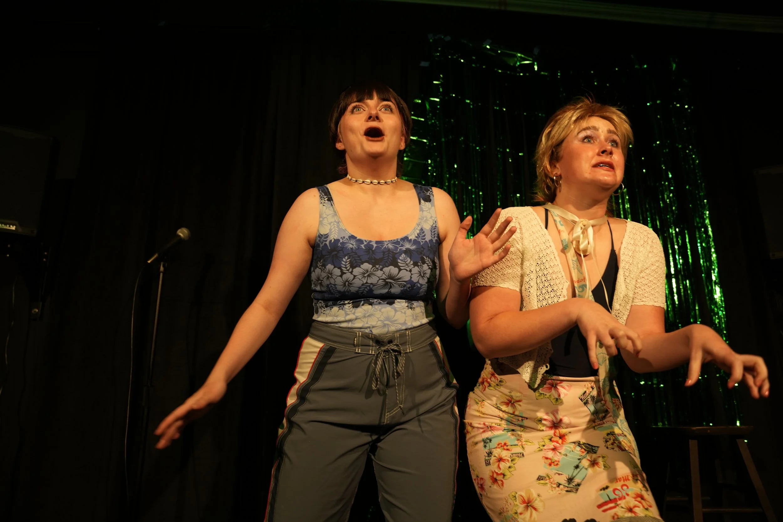 Two women standing on stage, performing with expressions of surprise or shock. One woman is wearing a floral tank top and gray shorts, the other is wearing a floral skirt and a cardigan. The background features dark curtains and green sparkly decorations.