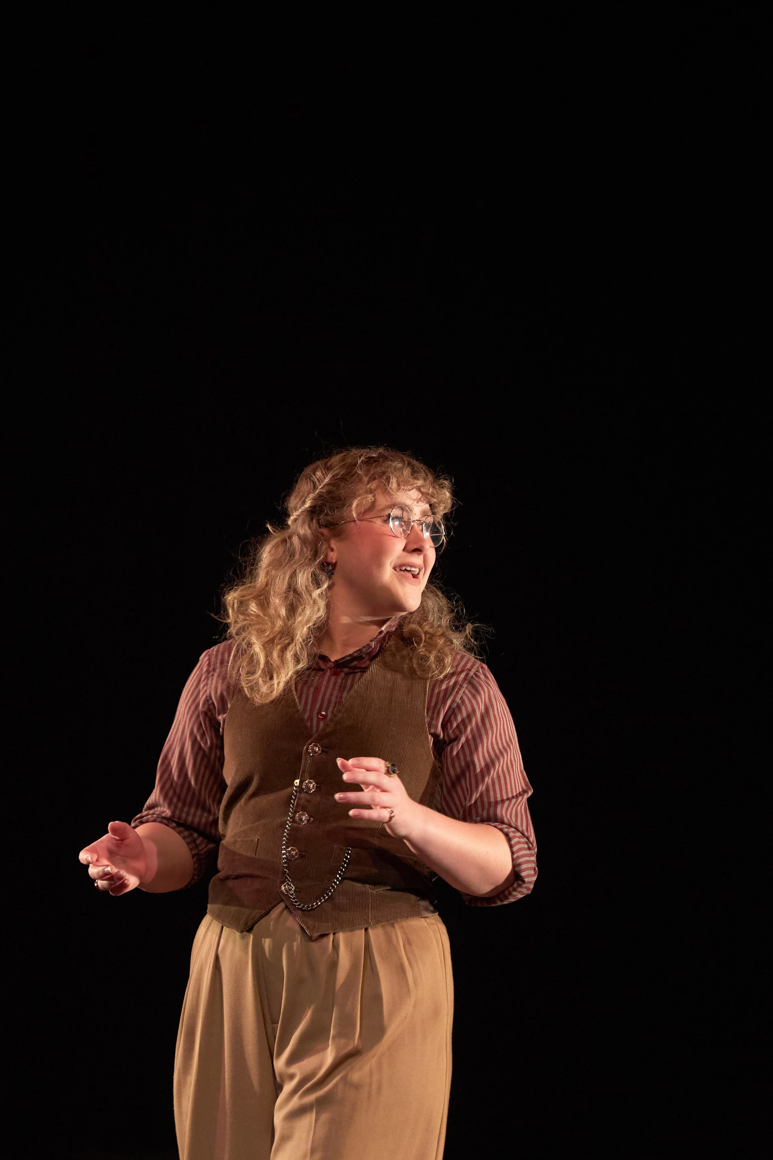 A woman with curly blonde hair, glasses, and a patterned shirt, smiling and gesturing with her hands against a black background.