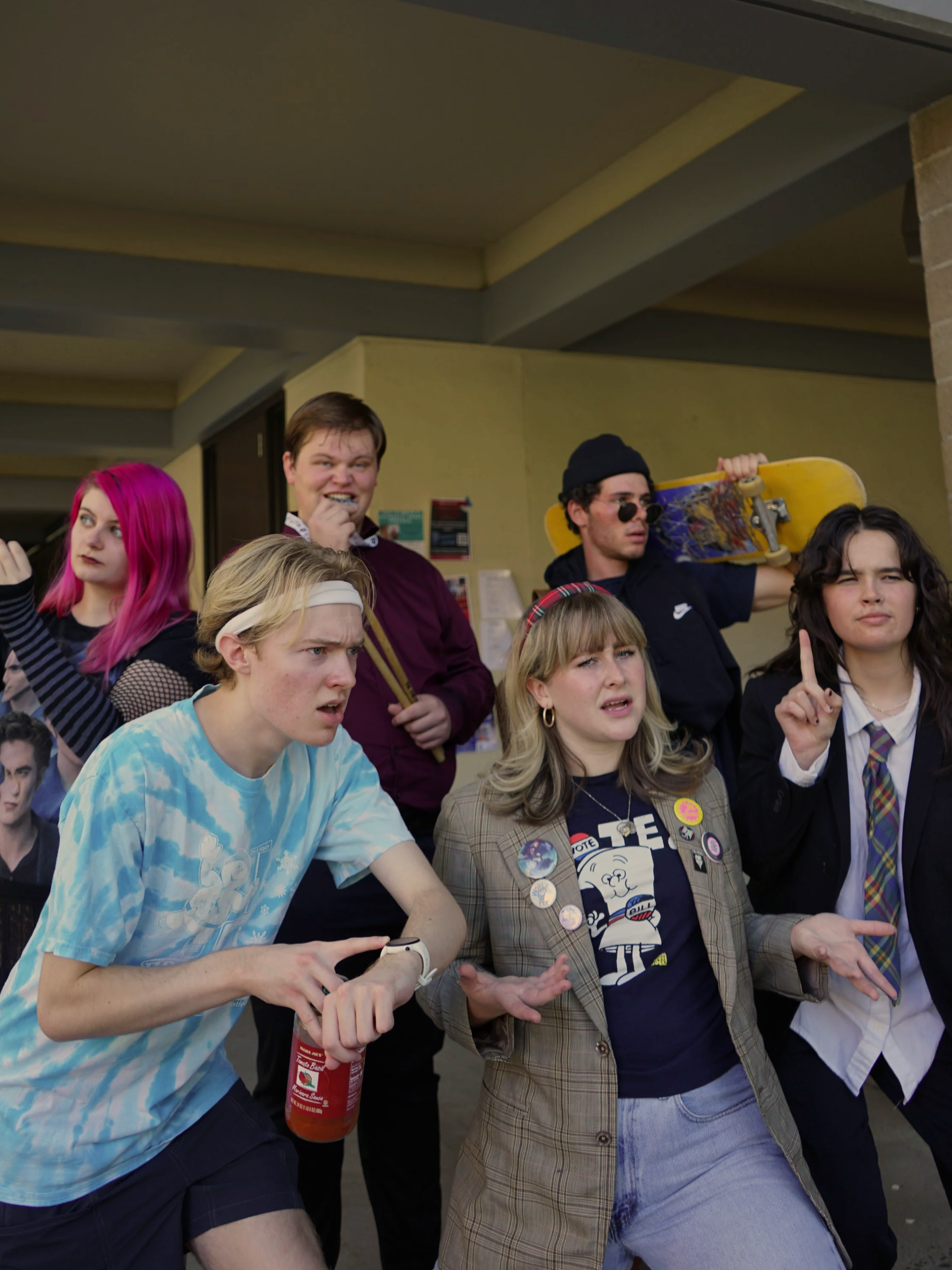 Group of young adults standing close together indoors, some with expressive facial gestures, one holding a skateboard, and another holding a bottle of ketchup.