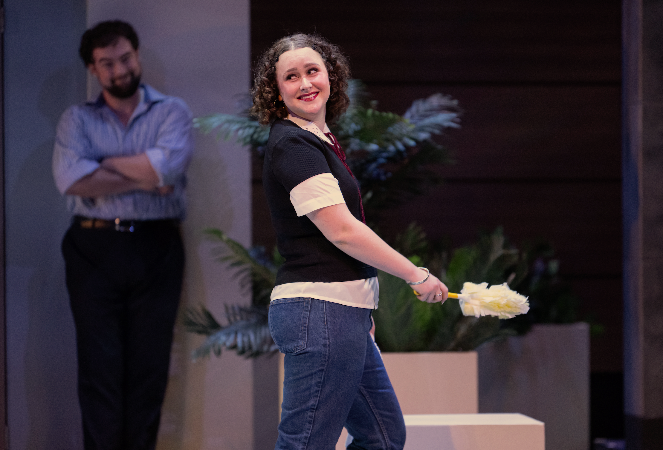 A woman with curly hair and wearing a black top with white cuffs holding a yellow-handled duster or feather duster. A man with dark hair and beard, wearing a blue striped shirt with rolled-up sleeves, stands in the background with his arms crossed, smiling and looking at her. The setting appears to be indoor with potted plants in the background.