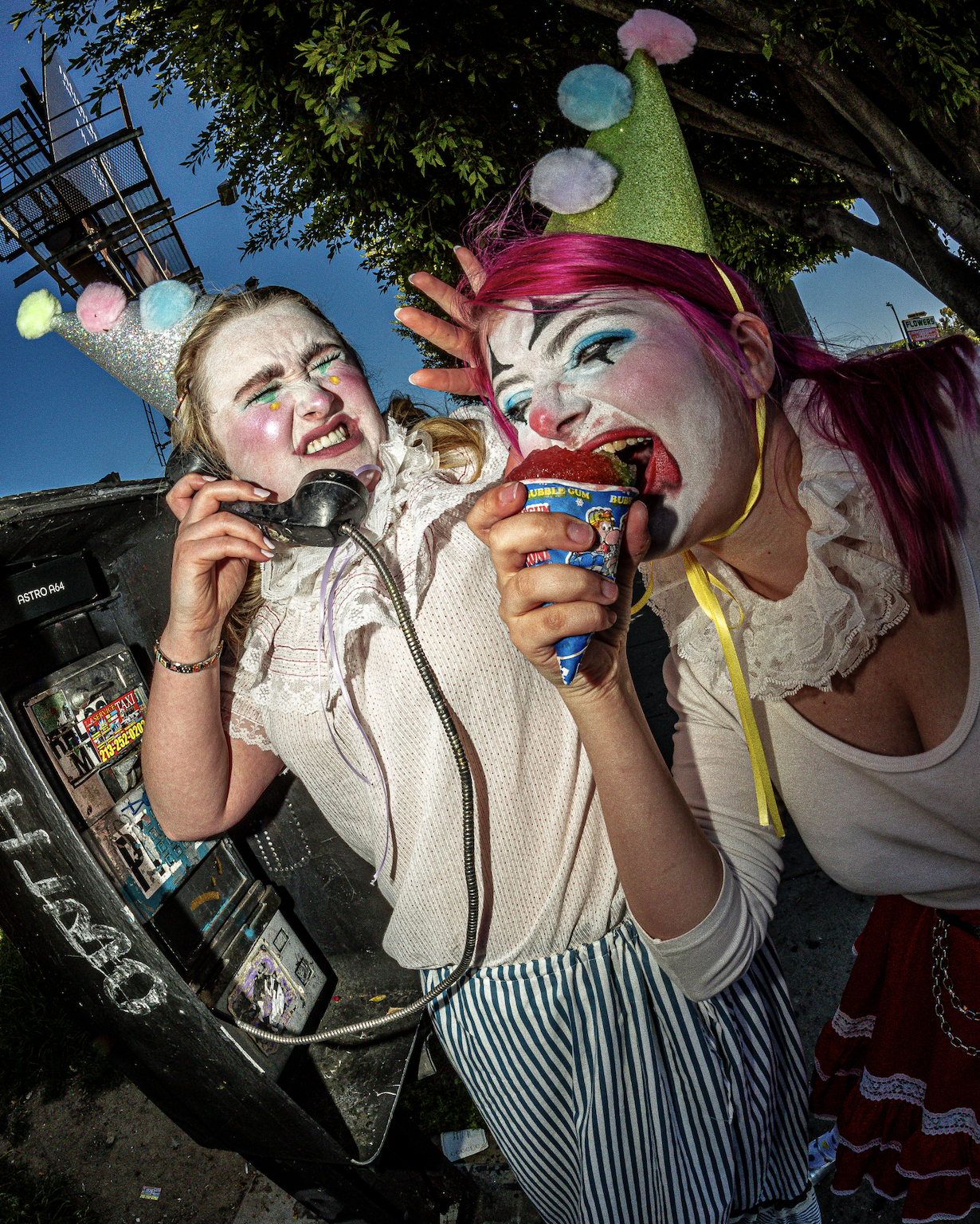 Two people dressed as clowns with colorful makeup and clothing, one wearing a party hat and the other with pink hair, sharing an ice cream cone outside near a tree.