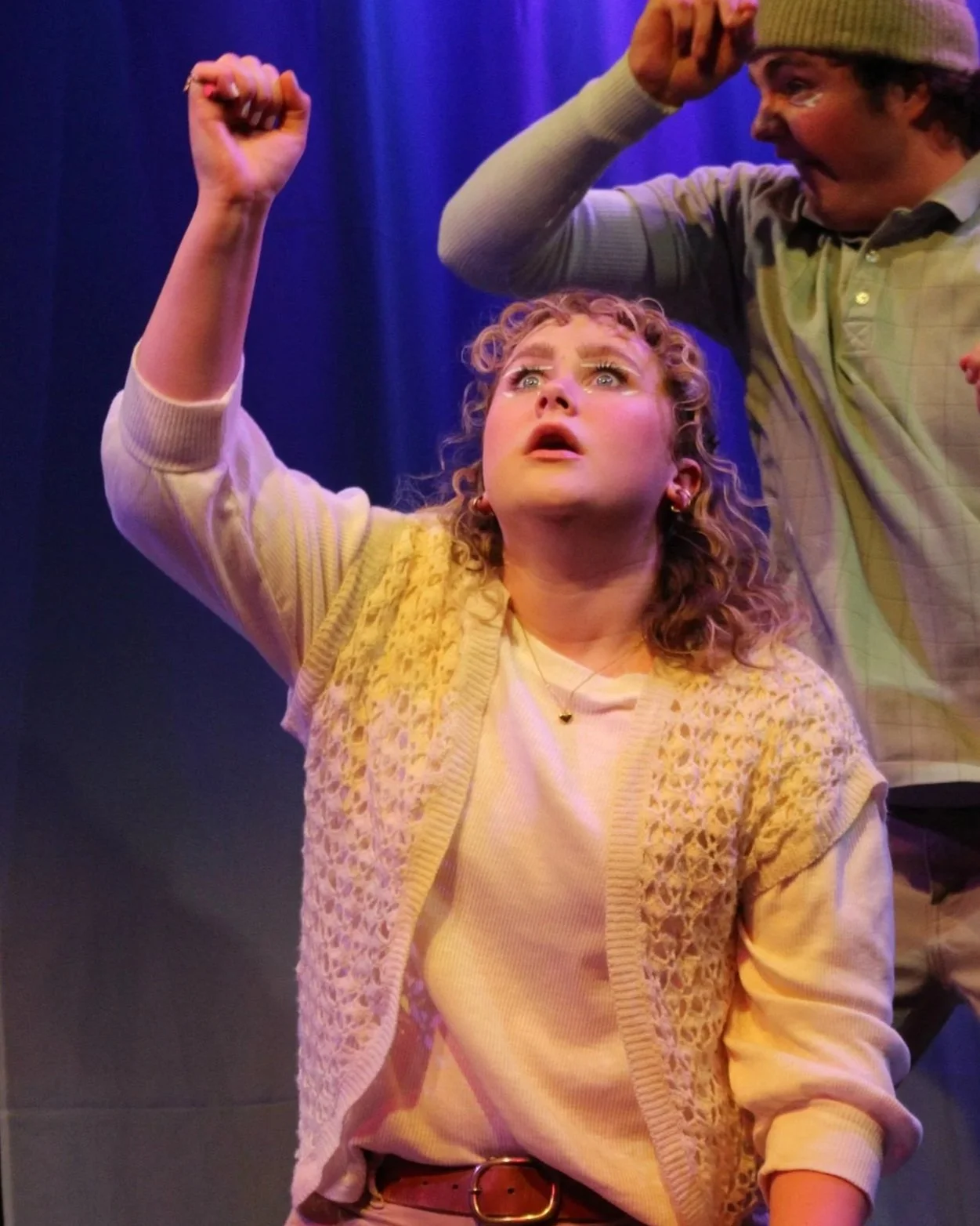 A young woman with curly hair and bright eyes looks upward with a determined expression, raising her right fist in the air. She is wearing a white top, a yellow patterned cardigan, and a gold necklace, with a dark background lit with colorful stage lighting.