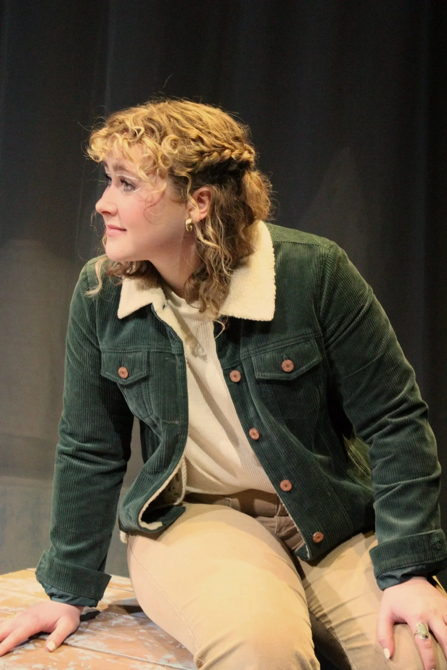 A woman with curly blonde hair in a braided style, wearing a green corduroy jacket with a shearling collar, sitting on a wooden surface against a black background.