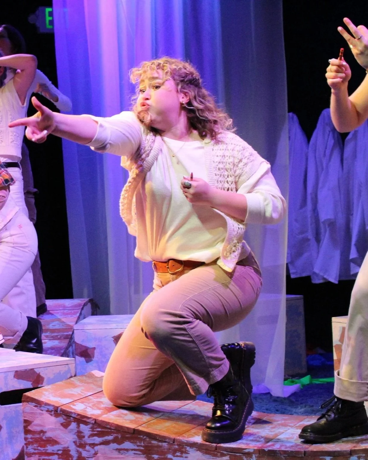 Person with curly hair wearing a cream-colored cardigan and pants, kneeling on one knee on stage during a theatrical performance, with background curtains and other actors visible.