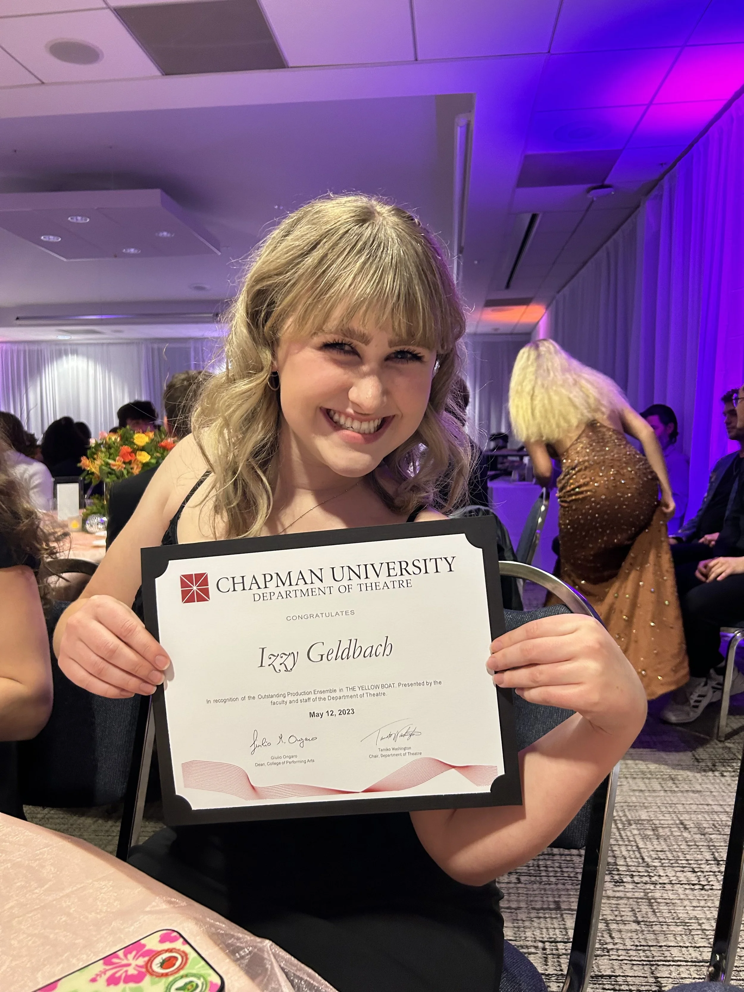 A young woman with blonde hair smiling and holding a diploma from Chapman University, Department of Theatre, dated May 12, 2023, at a celebration event with other people and colorful lighting in the background.