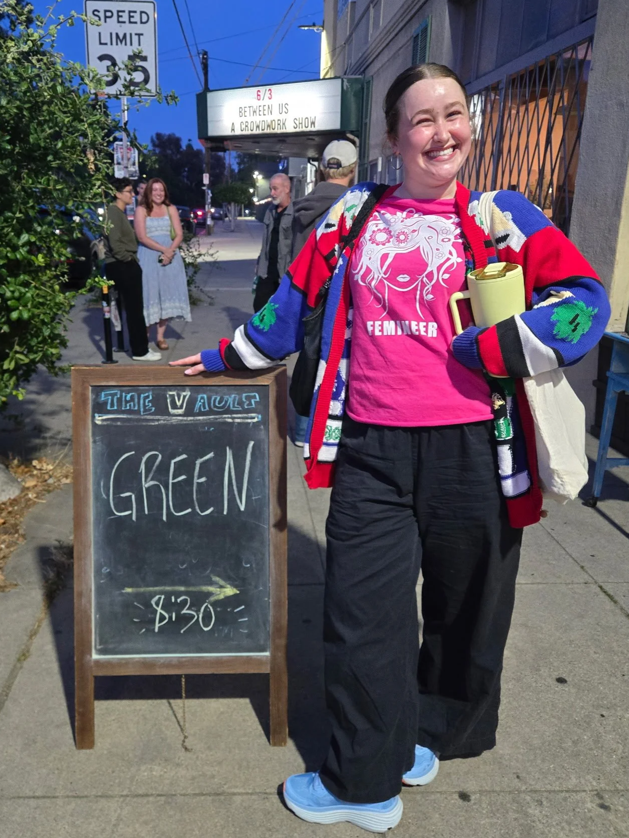 A young woman smiling and standing on a sidewalk outside a theater, holding a yellow mug. She is wearing a pink shirt with a white graphic of a woman's face and the word 'FEMMEHER,' a colorful cardigan, and black pants. A chalkboard sign next to her advertises a show called 'The Vals' at 8:30. Behind her, pedestrians are walking, and a marquee sign indicates a show called 'Between Us: A Crowdwork Show' at 6/3.
