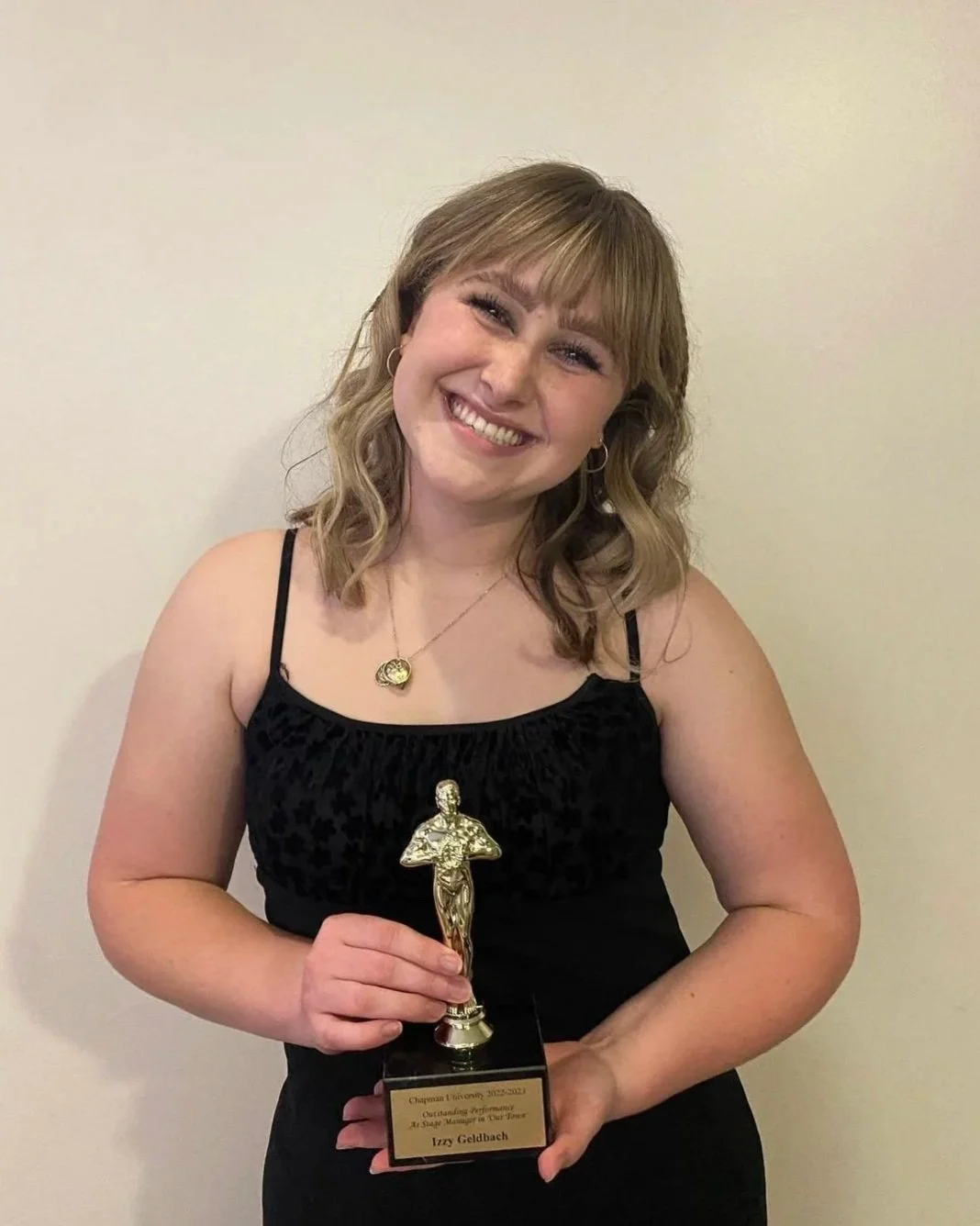 A young woman with shoulder-length blonde hair, smiling, holding an award trophy, wearing a black dress with thin straps and gold jewelry, standing against a plain light-colored wall.