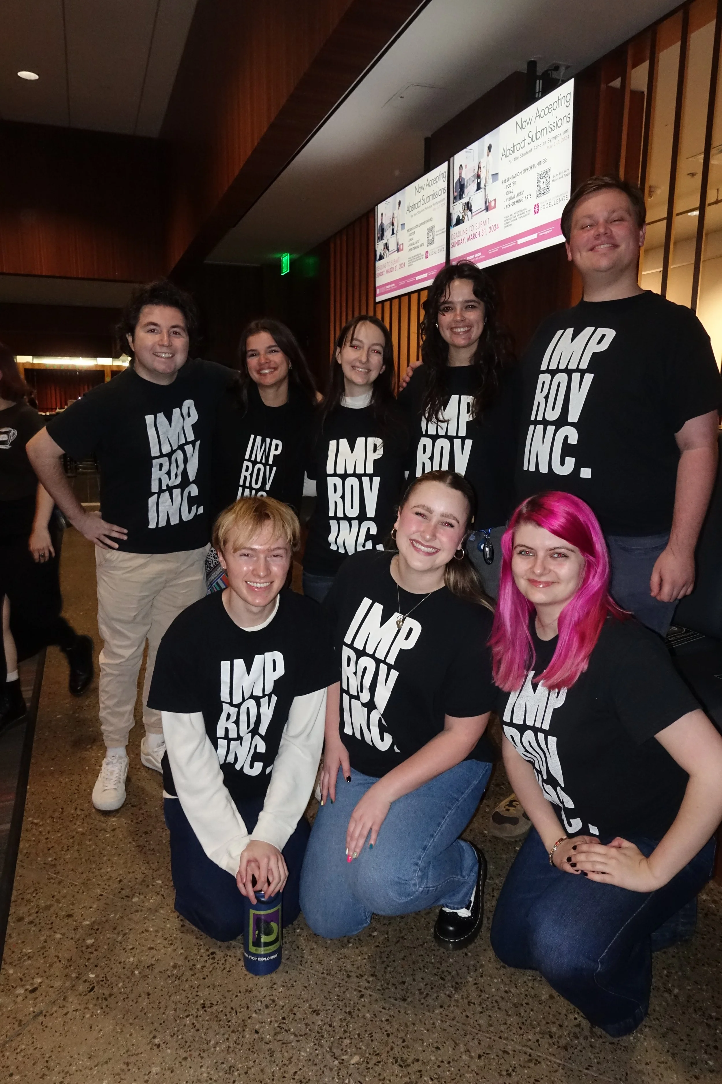 Group of young people smiling, wearing black t-shirts with 'IMP ROV INC.' printed on them, in an indoor setting with a large digital screen in the background displaying acceptance and submission information.
