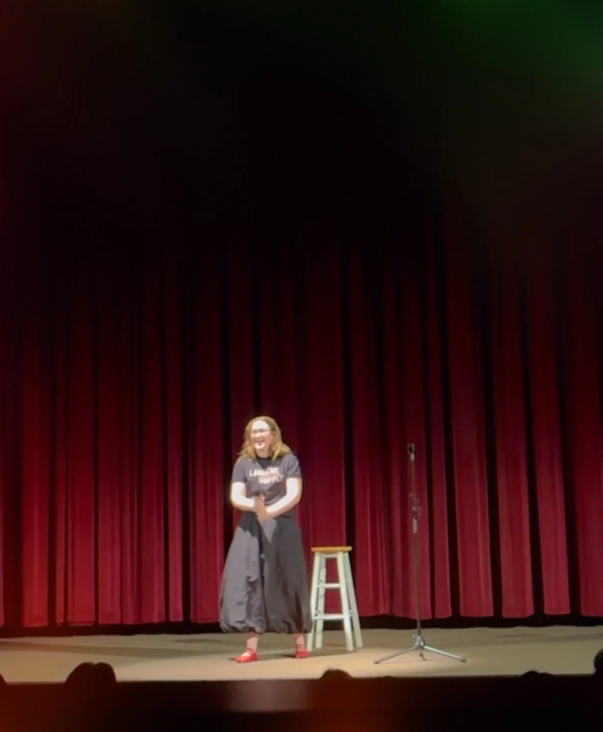 A woman stands on a stage in front of a red curtain, holding a microphone with a stool nearby. She is smiling and appears to be performing or speaking.